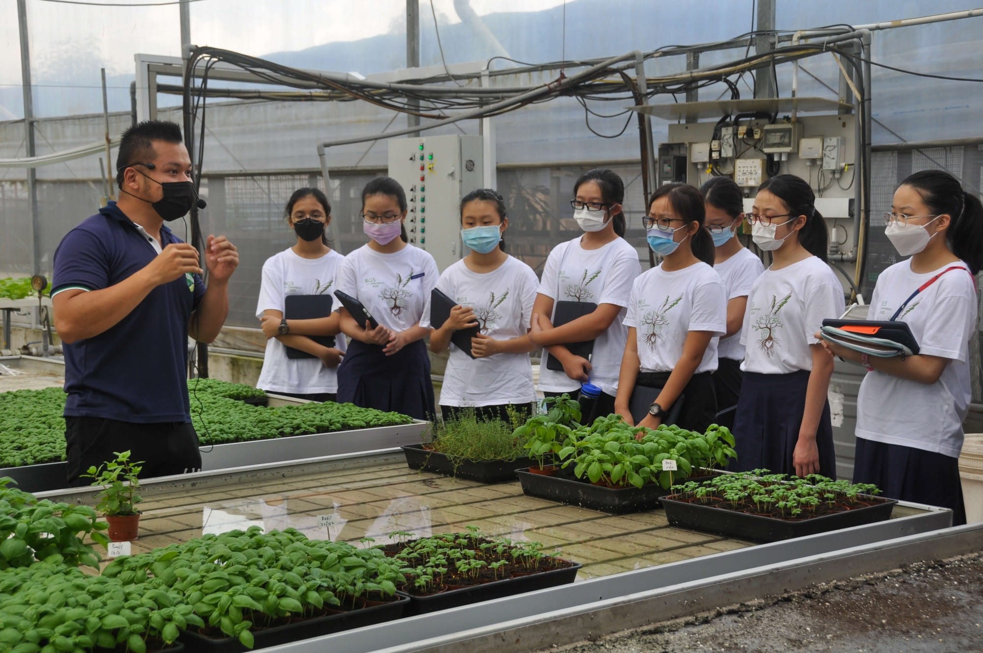 A masked instructor speaks to a group of masked students in white shirts inside a greenhouse with plant trays.