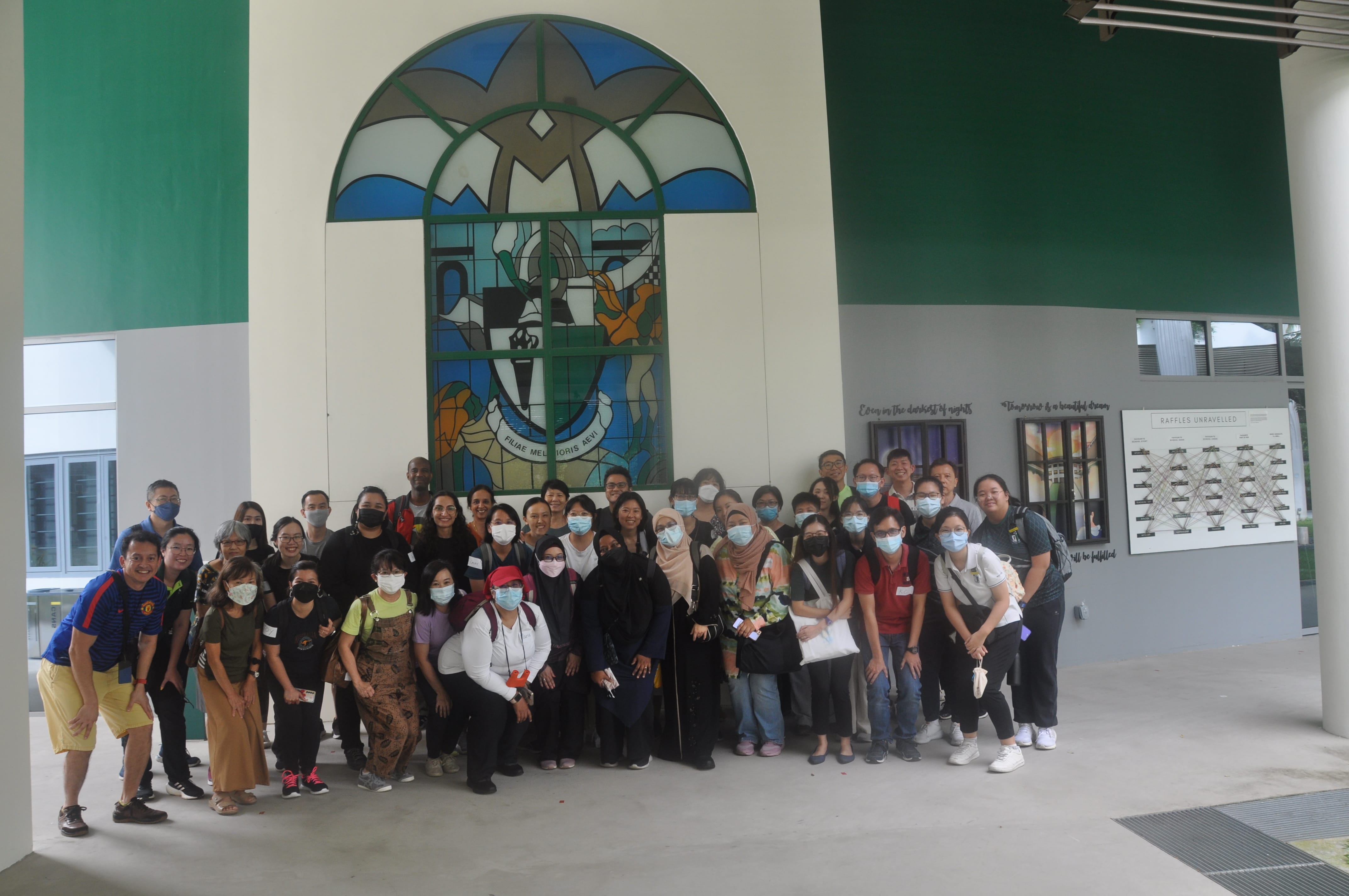 Group of people wearing masks posing under a large arched stained-glass window in a covered walkway