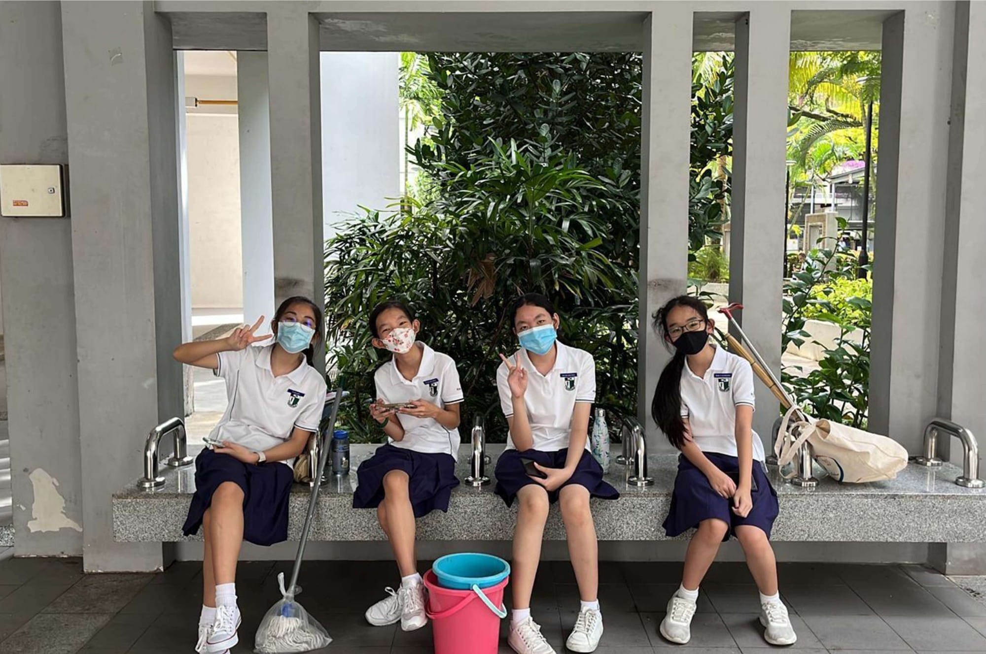 Four masked schoolgirls in white uniforms sit on a bench outdoors with cleaning supplies, posing with peace signs.