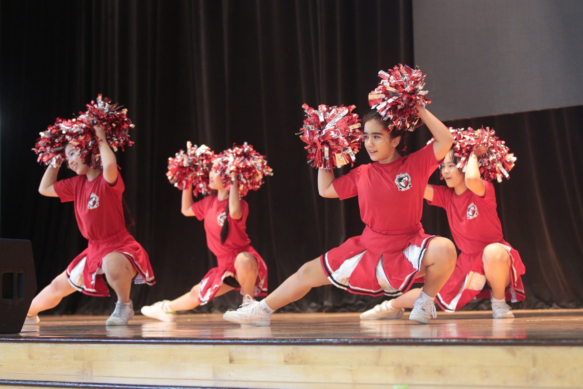 Four young cheerleaders in red uniforms perform a kneeling routine with red pom-poms on a wooden stage.