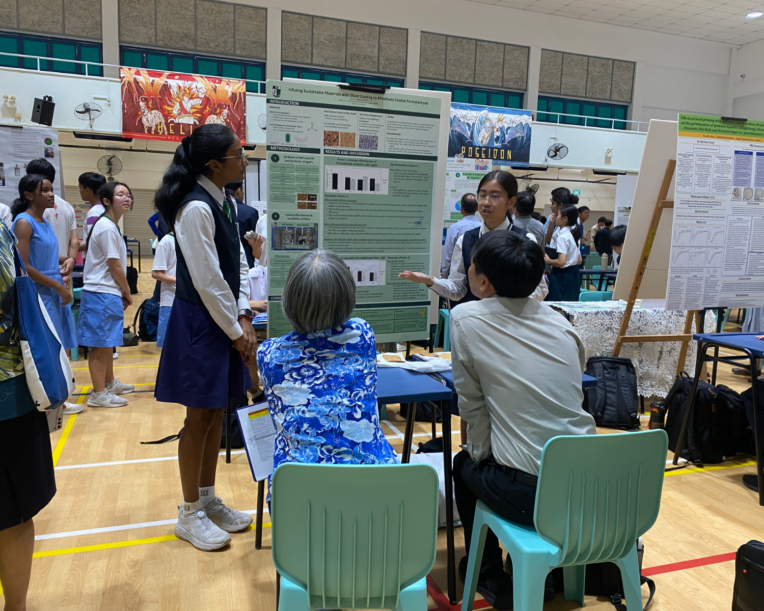 Students present a green science poster to seated judges at a busy school fair in a gymnasium.