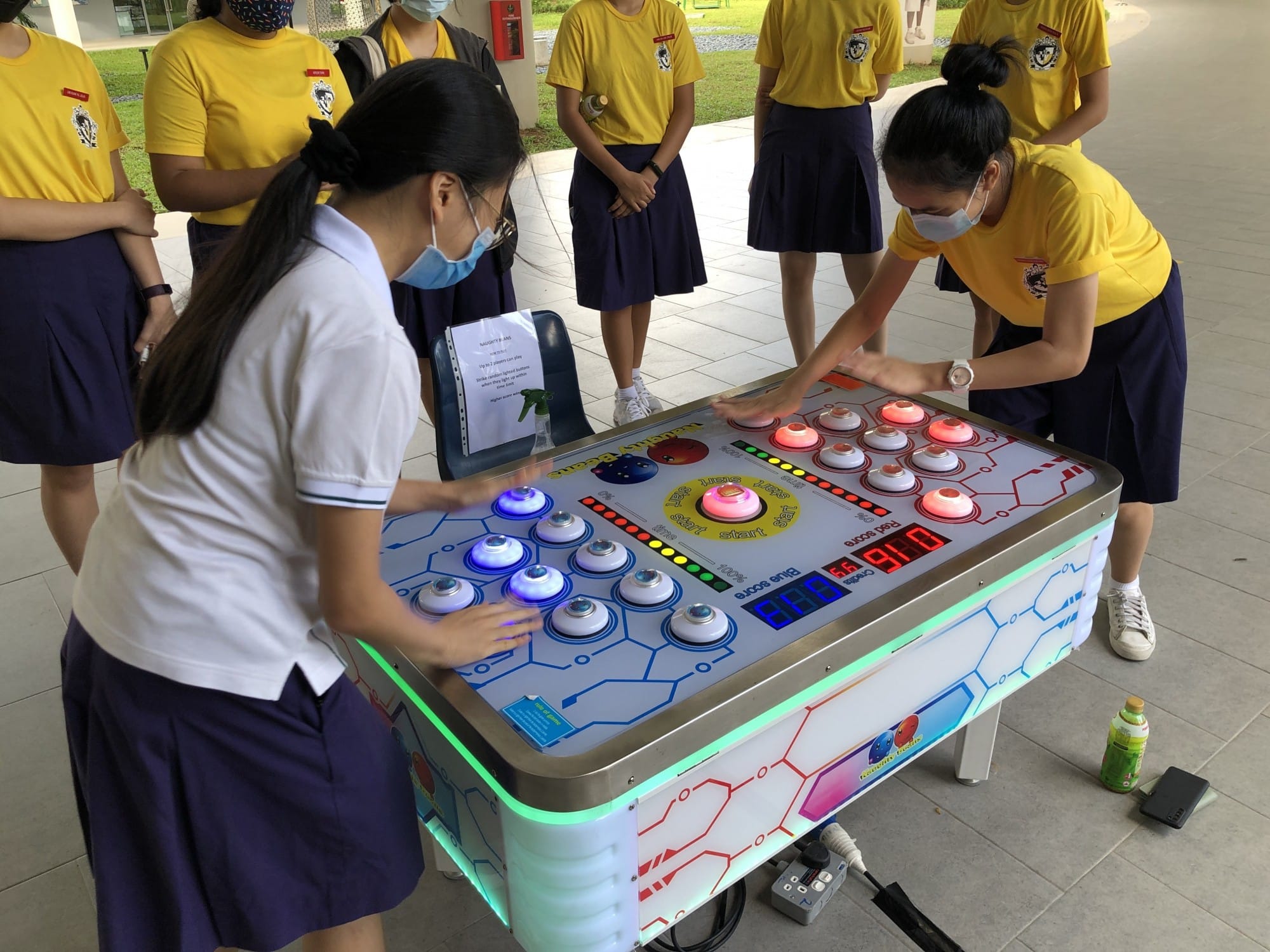 Two masked students play a lit button arcade game while classmates in yellow shirts watch outdoors.