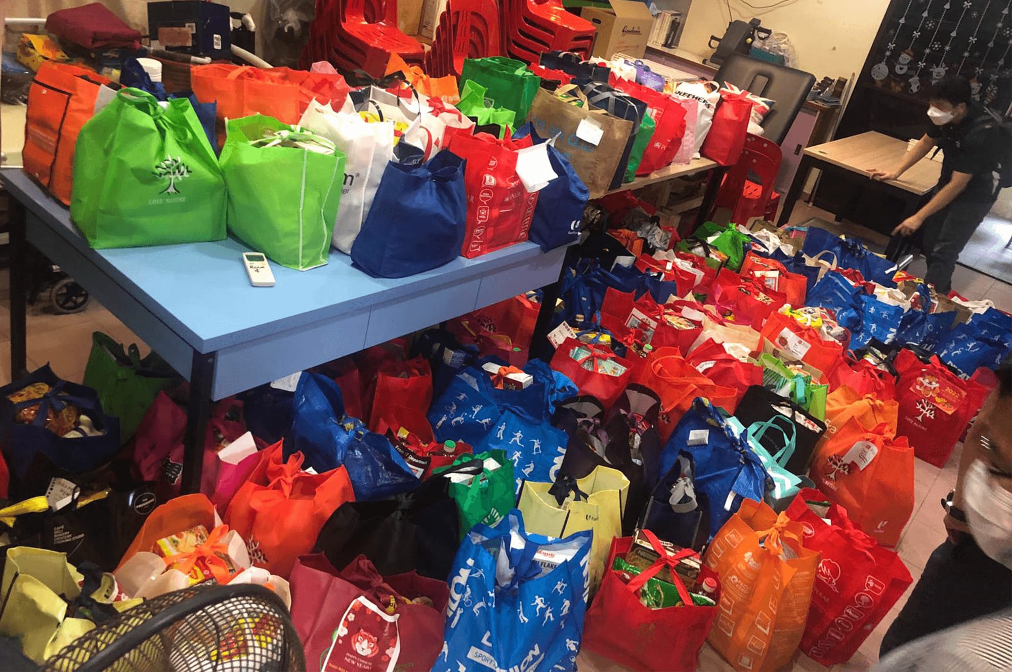 Dozens of colorful reusable bags filled with goods cover a floor and table, with a masked person standing nearby.