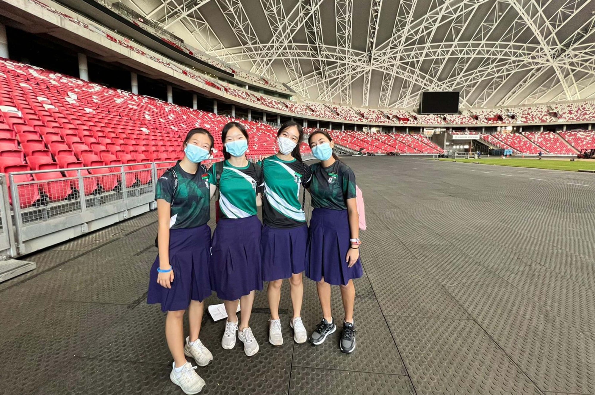 Four girls in green uniforms and blue skirts wearing face masks pose together inside a large empty stadium with red seats.