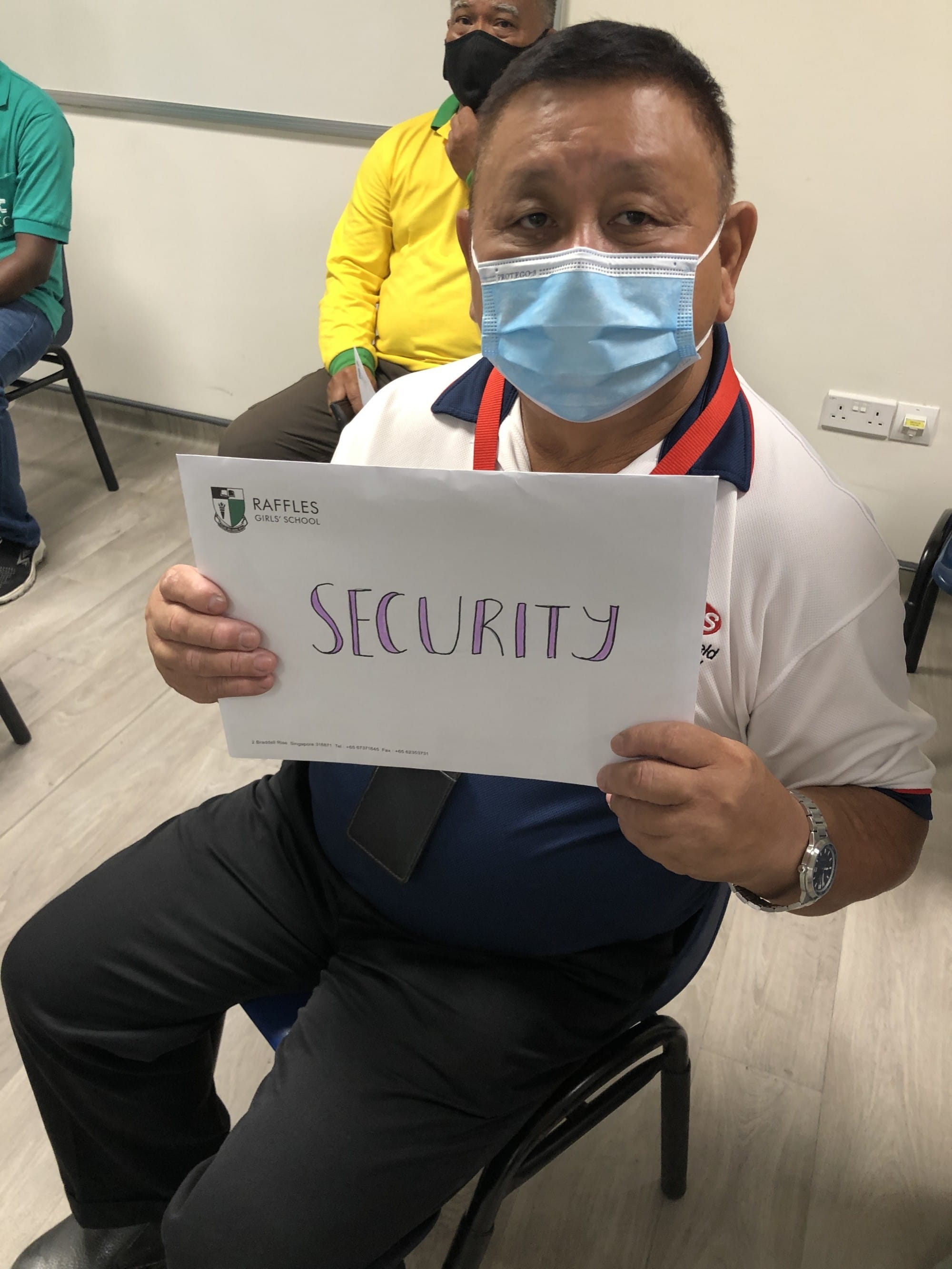 Man wearing a blue face mask holds a Raffles Girls School paper sign reading "SECURITY" in purple letters.