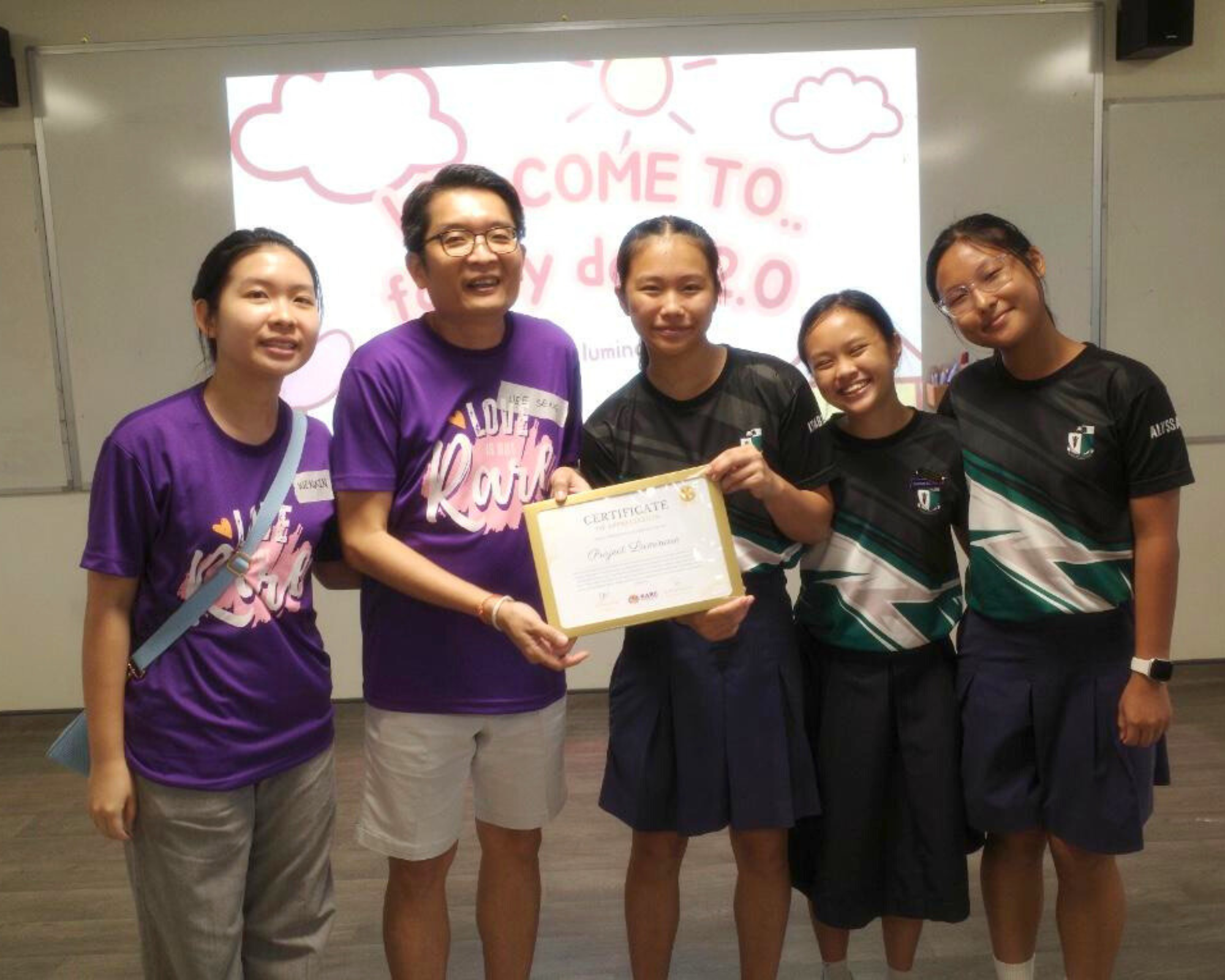 Five people smile while holding a certificate in front of a projected screen reading "Come To."
