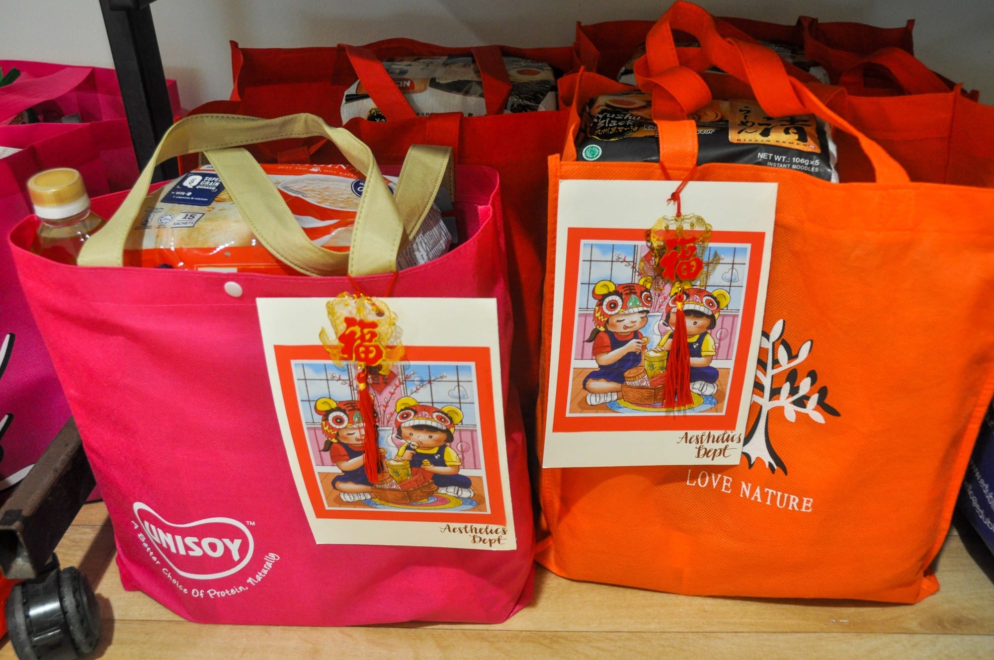 Red Unisoy and orange Love Nature tote bags with Chinese New Year cartoon children artwork, filled with food items on a shelf.
