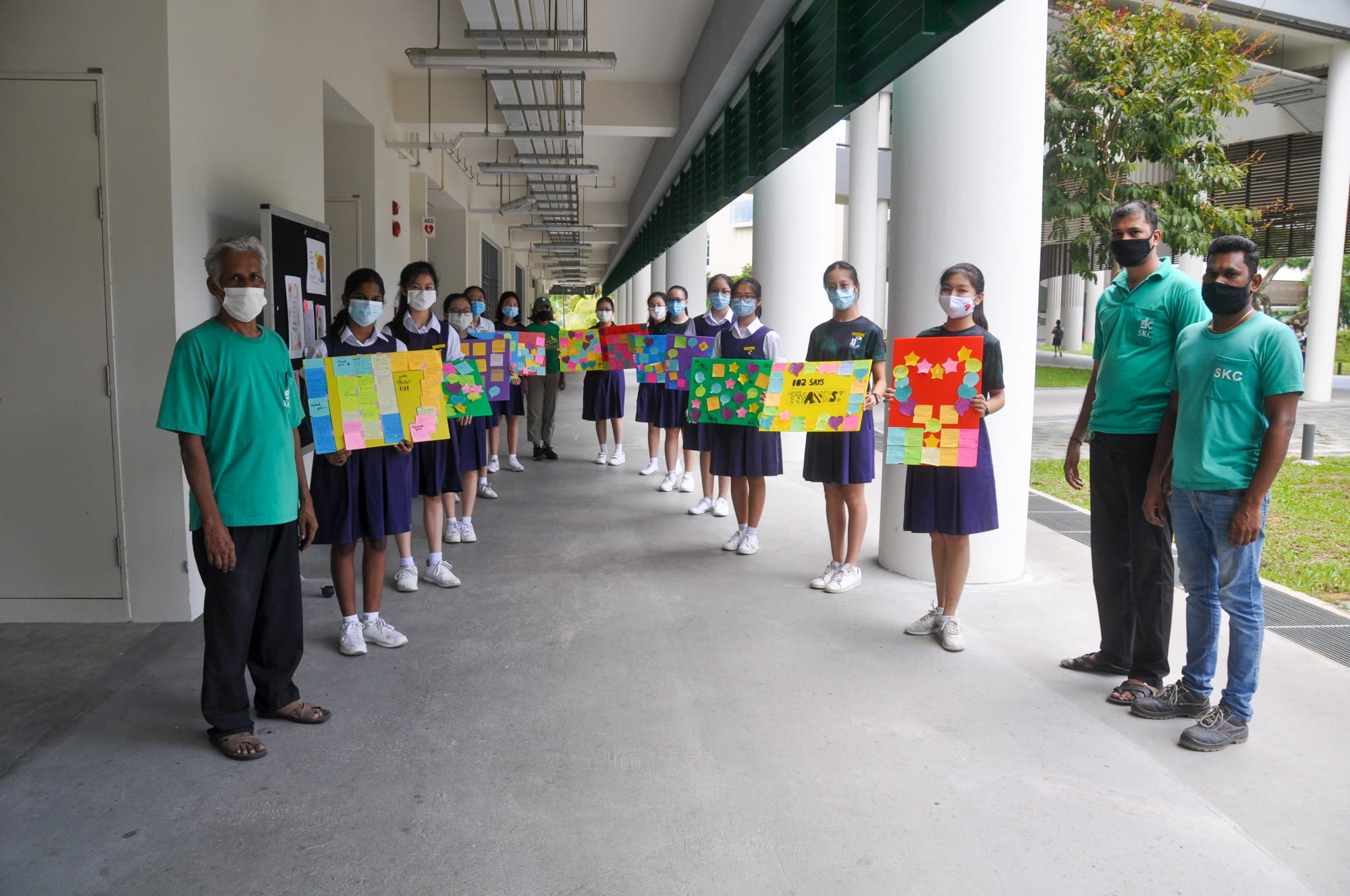 Students in purple uniforms and masked adults hold colorful hand-made posters in a school corridor.