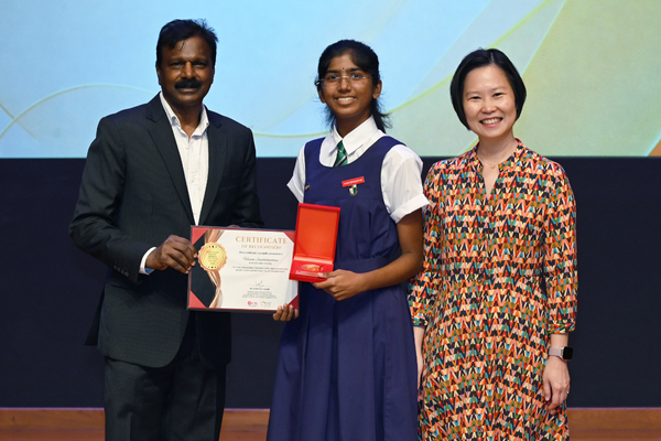 A man holds a certificate while a young girl in school uniform holds a red box, standing beside a woman in a patterned dress.