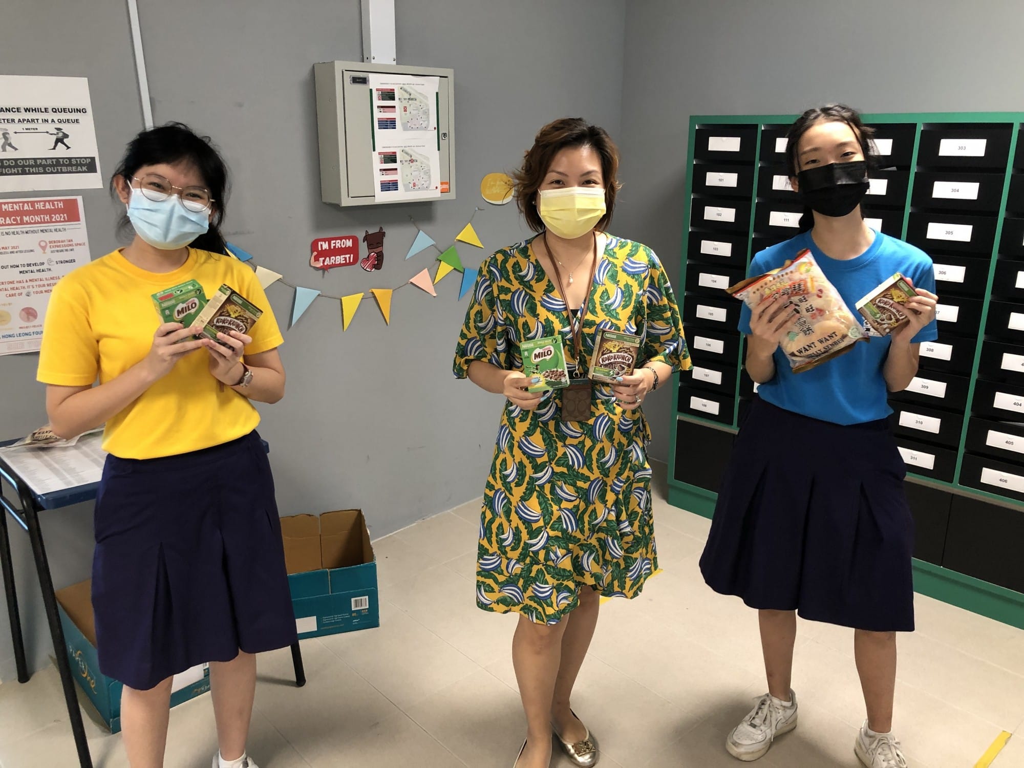 Three masked women stand indoors holding Milo and snack packages, with numbered drawers and posters on the wall behind them.