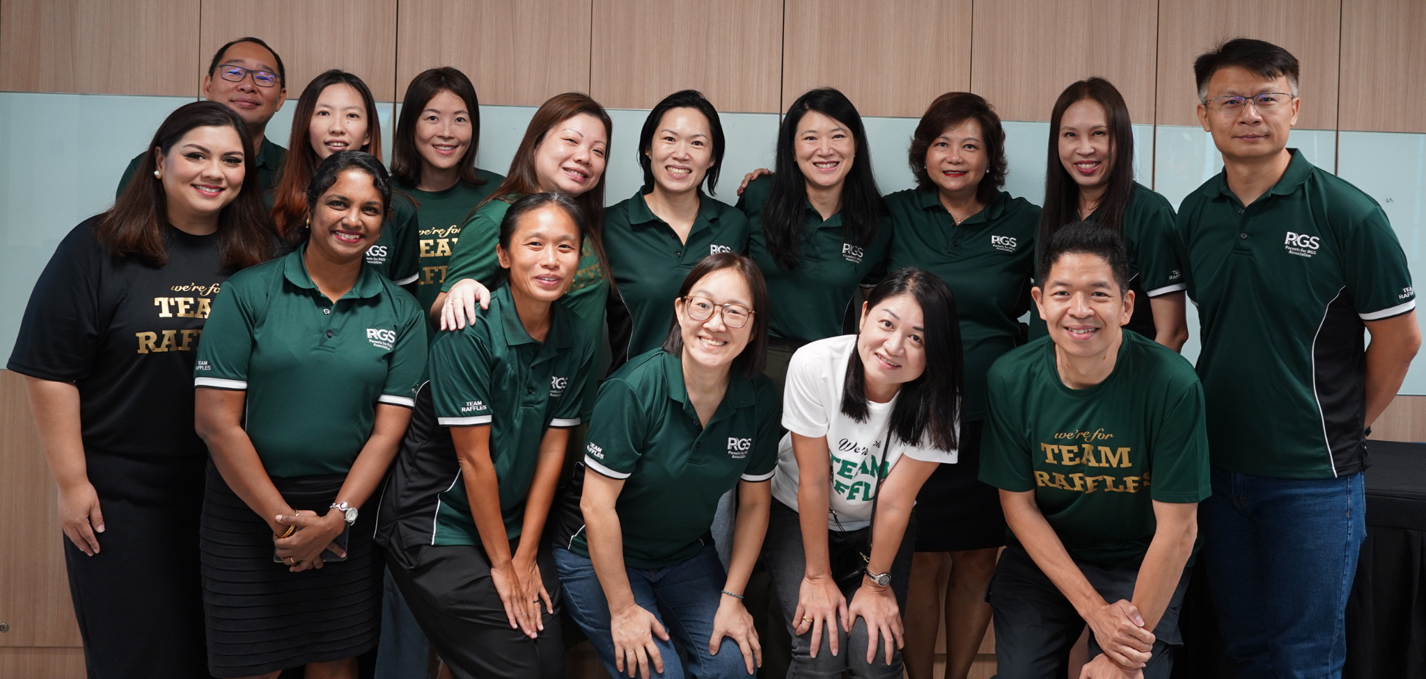Group of about 14 smiling adults in dark green RGS Team Raffles shirts posing together indoors.