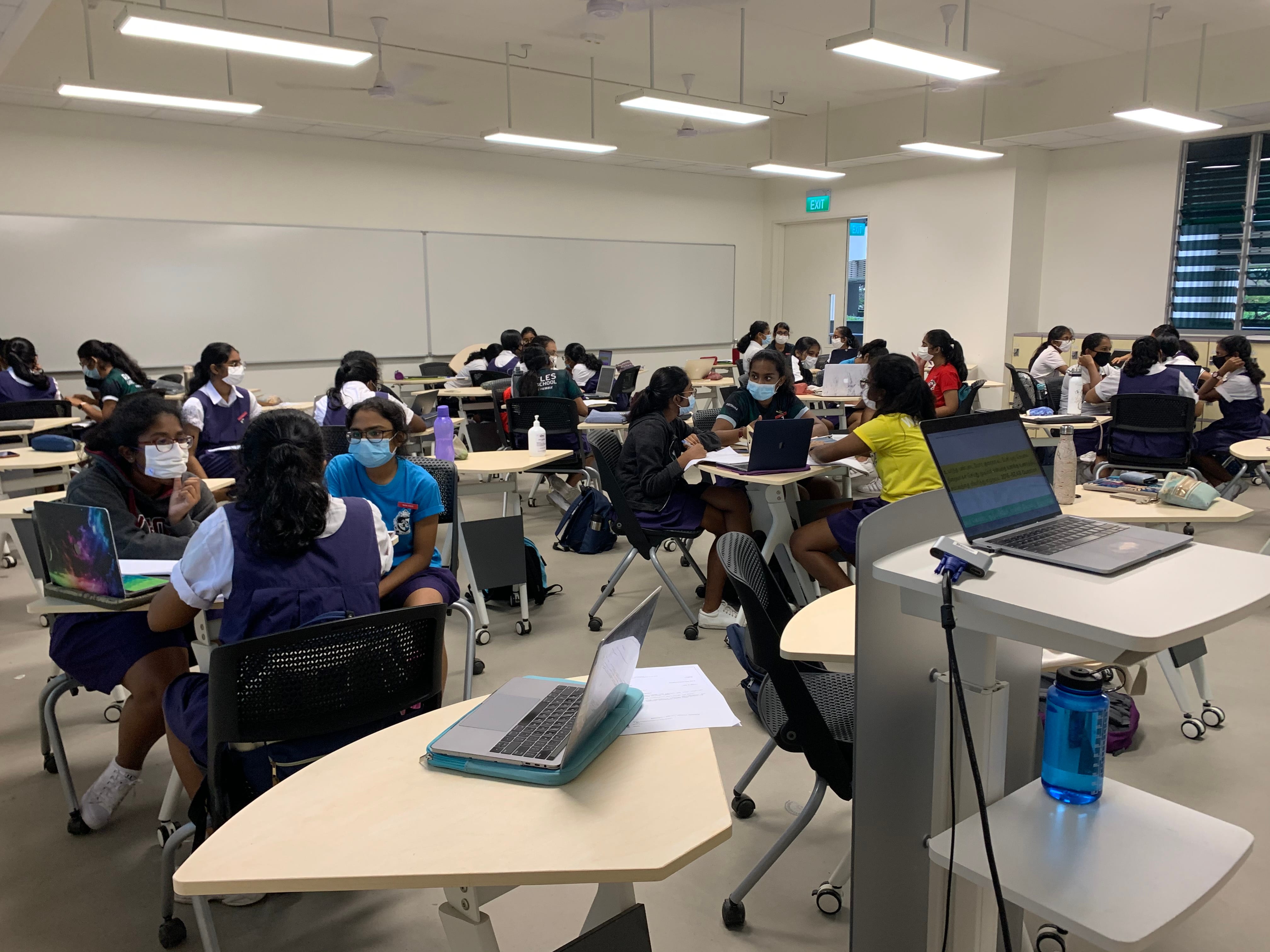 Students in school uniforms wearing masks sit at cluster desks with laptops in a bright modern classroom.