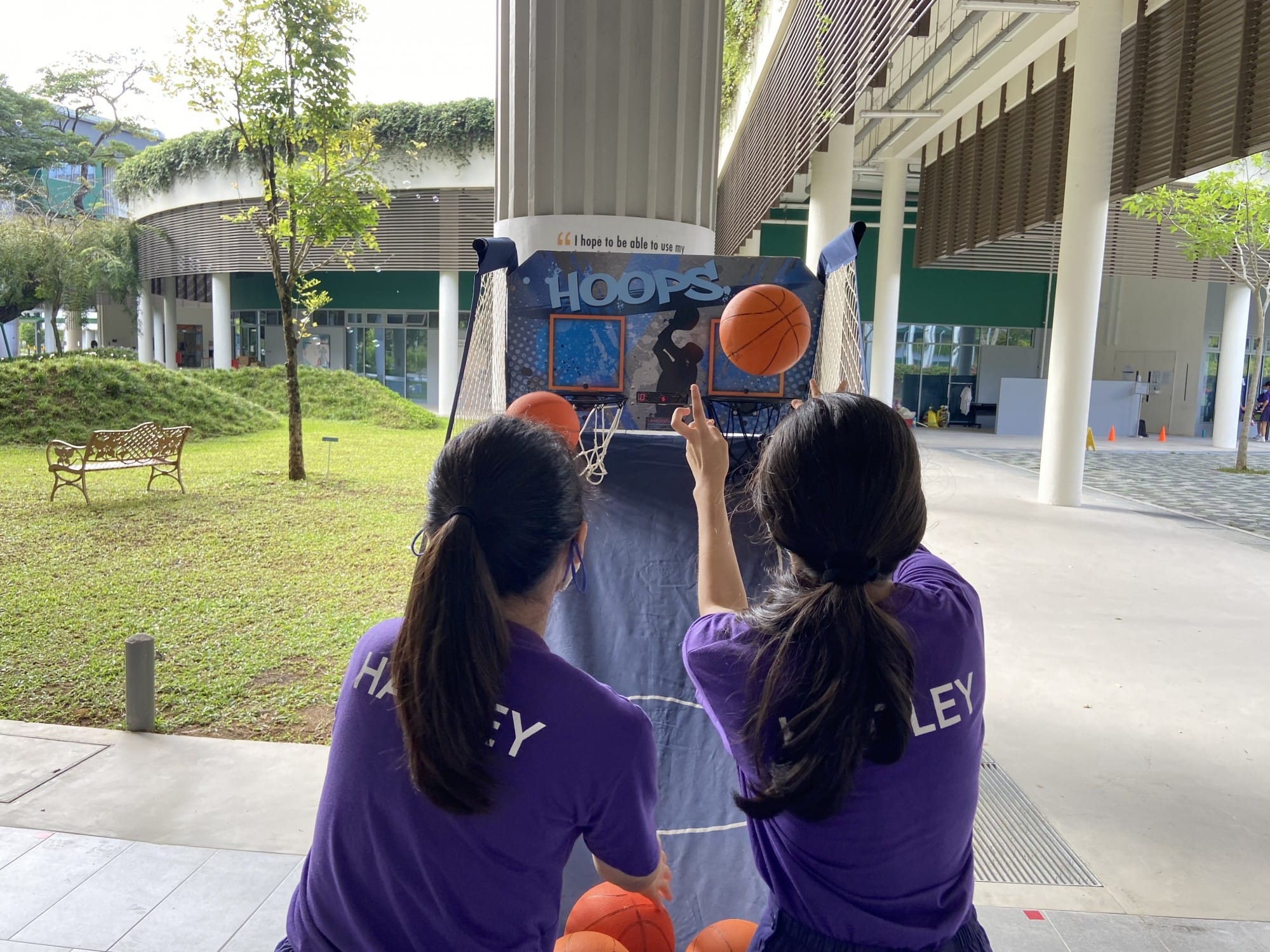 Two girls in purple shirts play an arcade basketball hoops game outdoors near a modern building.