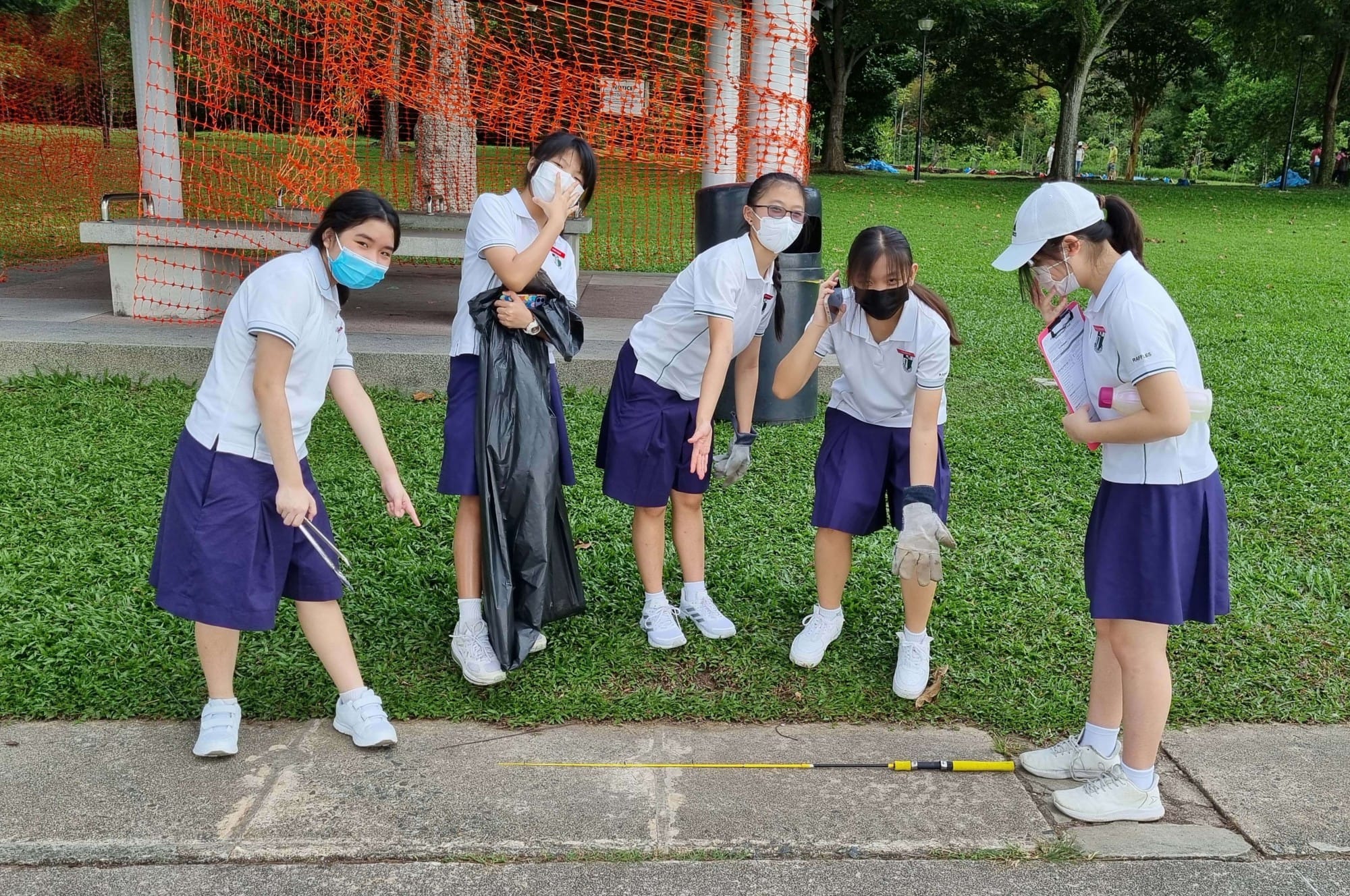 Five masked students in white uniforms collect litter in a park using bags and a grabber tool.