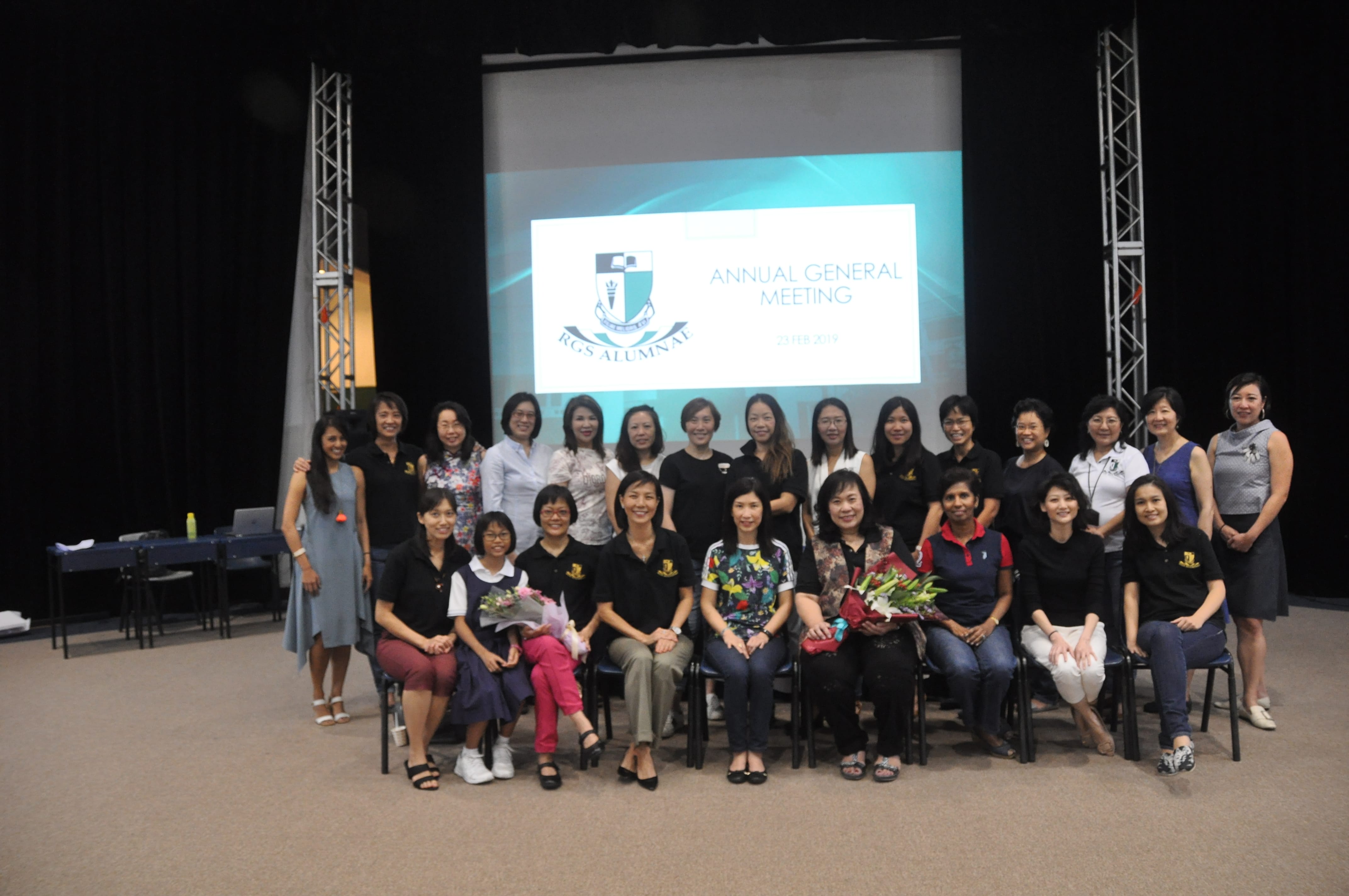 Group of women posing on stage under a screen reading Annual General Meeting, with some holding bouquets.