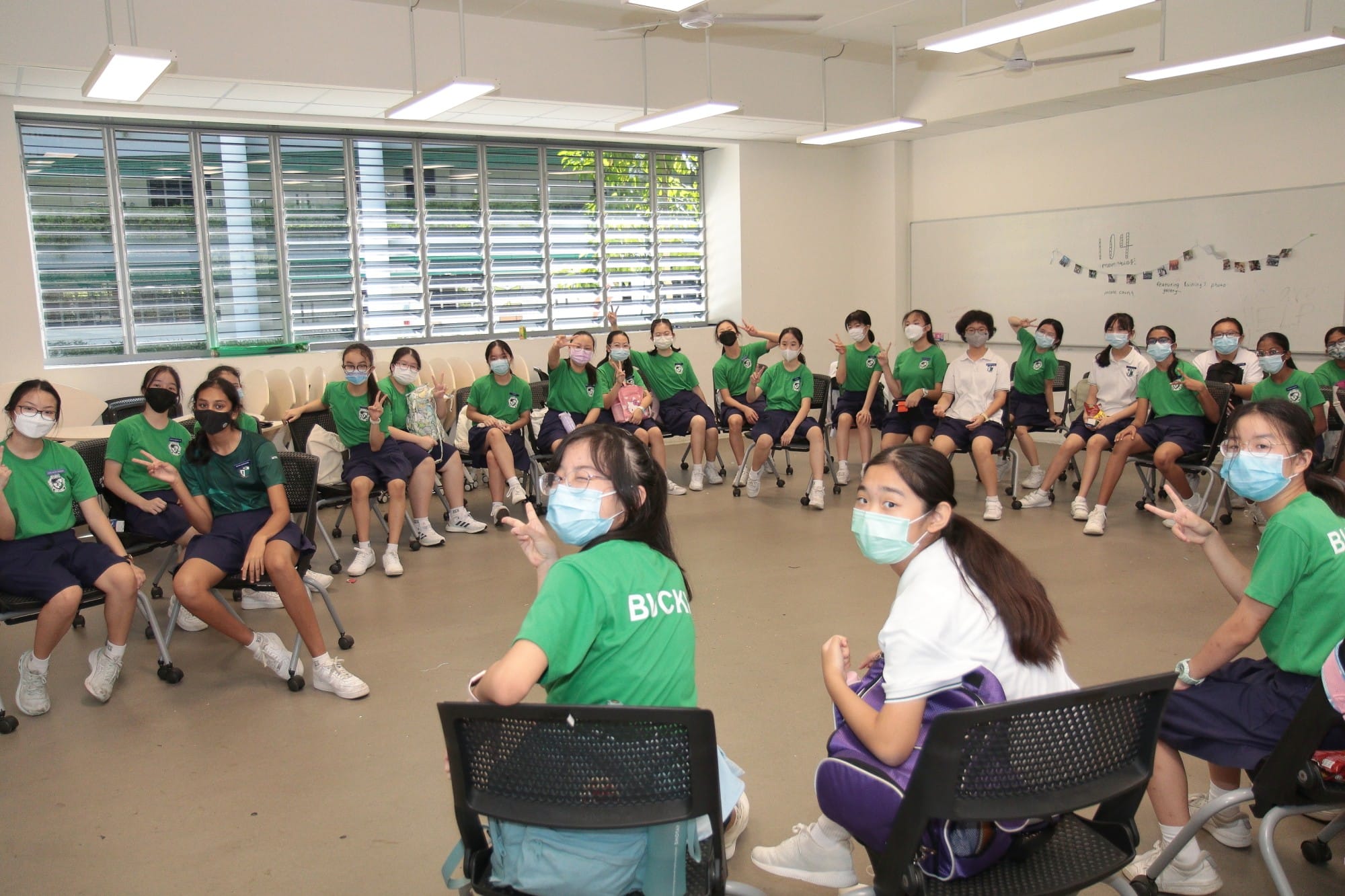 A large group of students in green uniforms and face masks sit in a circle in a bright classroom, many making peace signs.