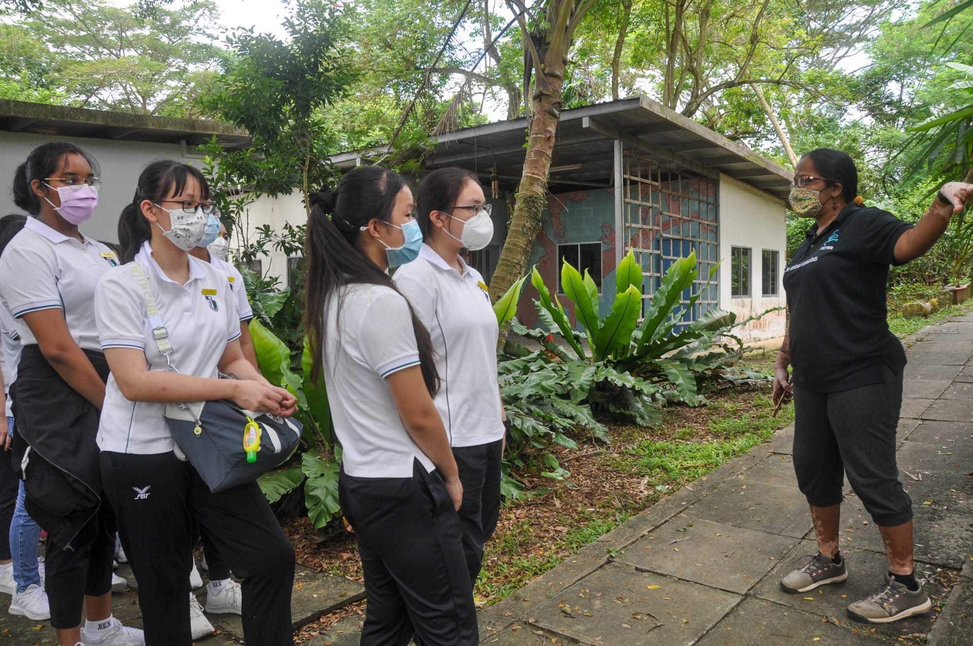 A masked instructor in black gestures while speaking to a group of masked students in white uniforms outdoors near a building.
