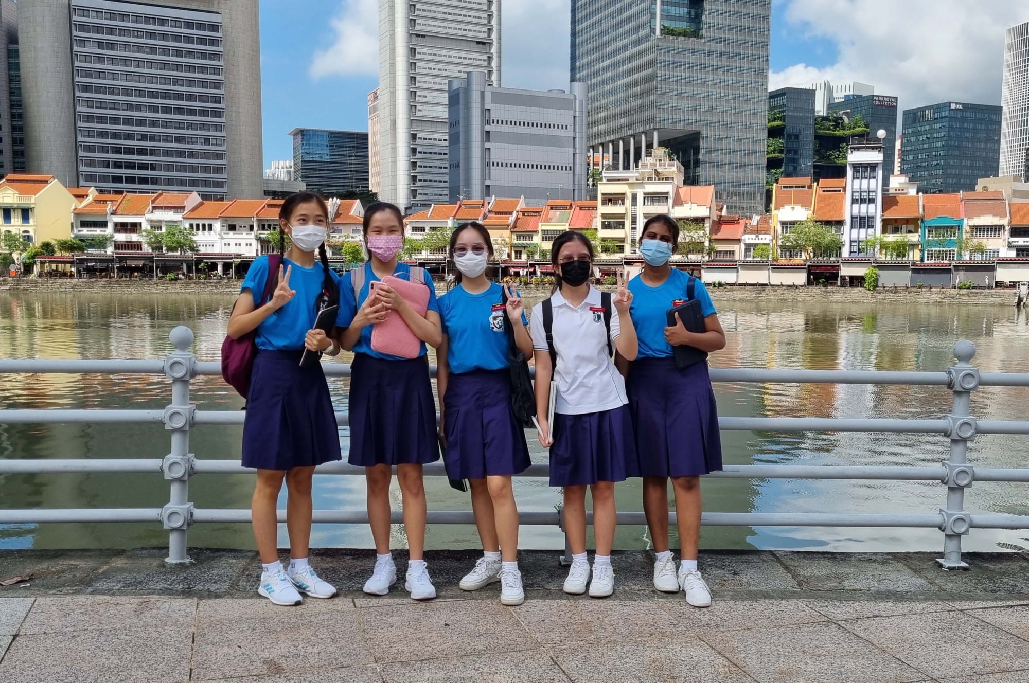 Five masked schoolgirls in blue uniforms pose by a river railing with Singapore city buildings in the background.