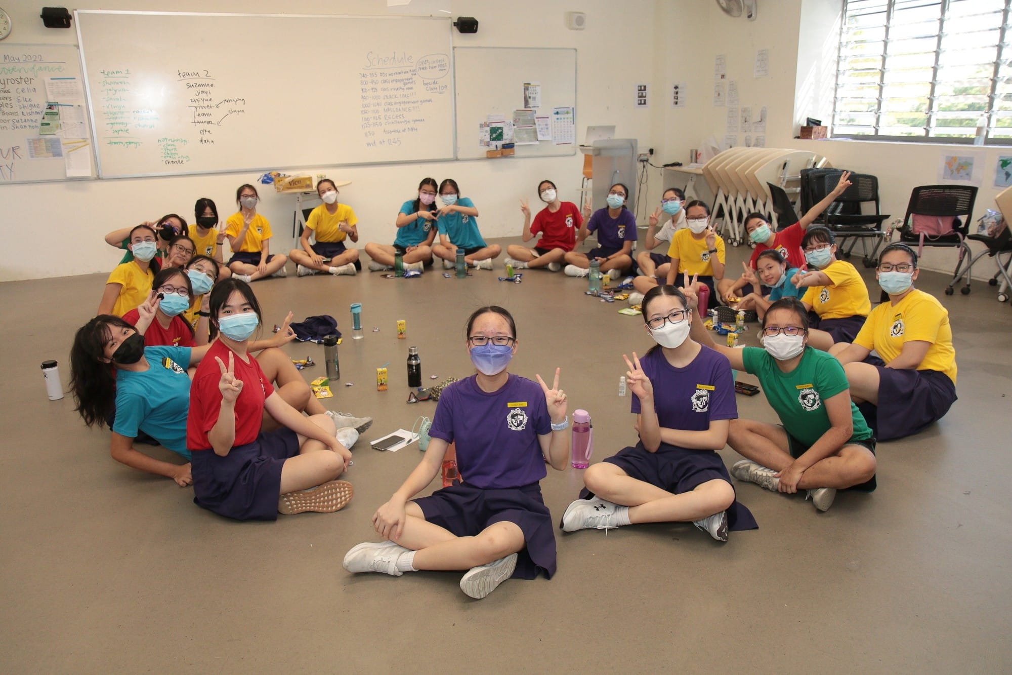 Students in colorful uniforms and face masks sit in a circle on a classroom floor, making peace signs.