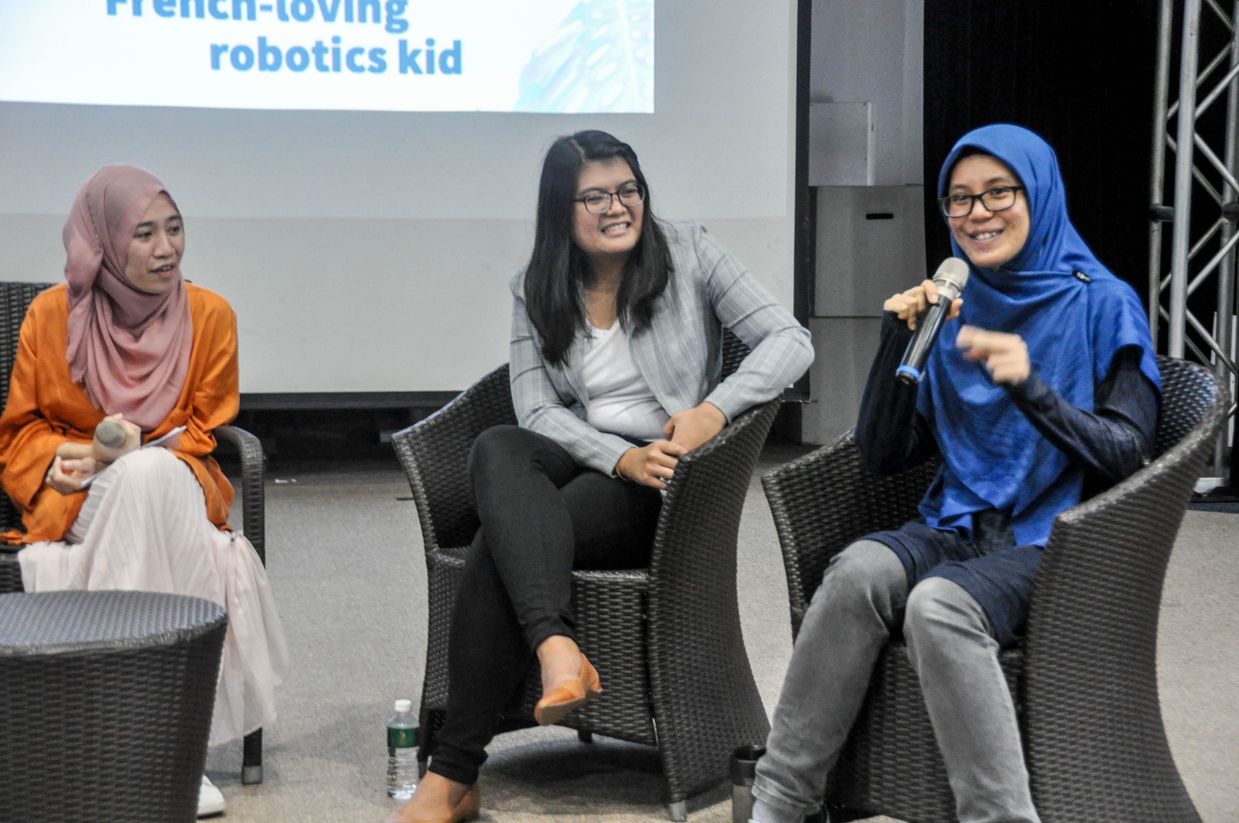 Three women sit in chairs on a stage, two wearing hijabs, one holding a microphone, smiling during a panel event.