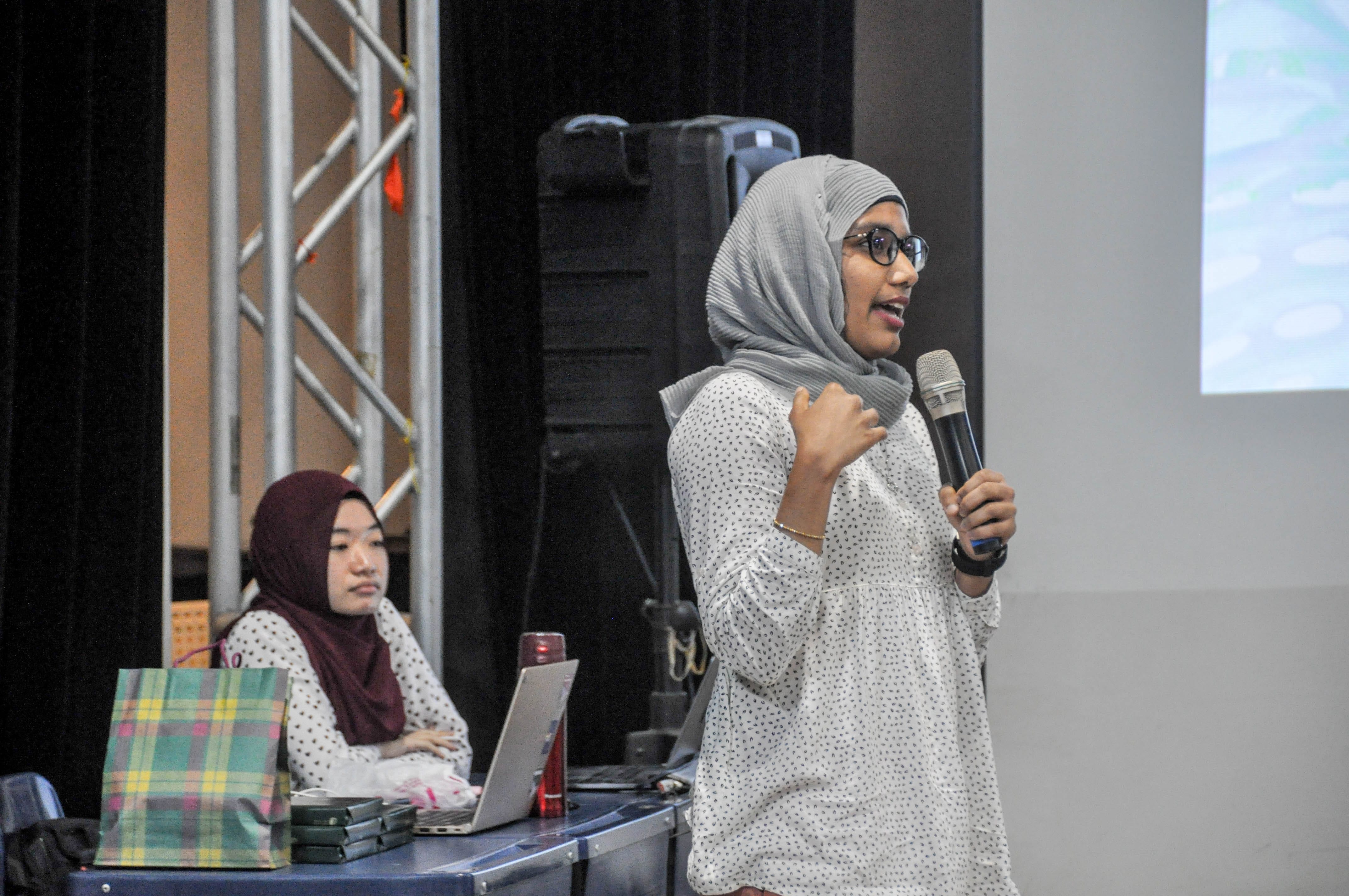 A woman in a gray hijab and glasses speaks into a microphone while another woman in a maroon hijab sits at a table with a laptop.