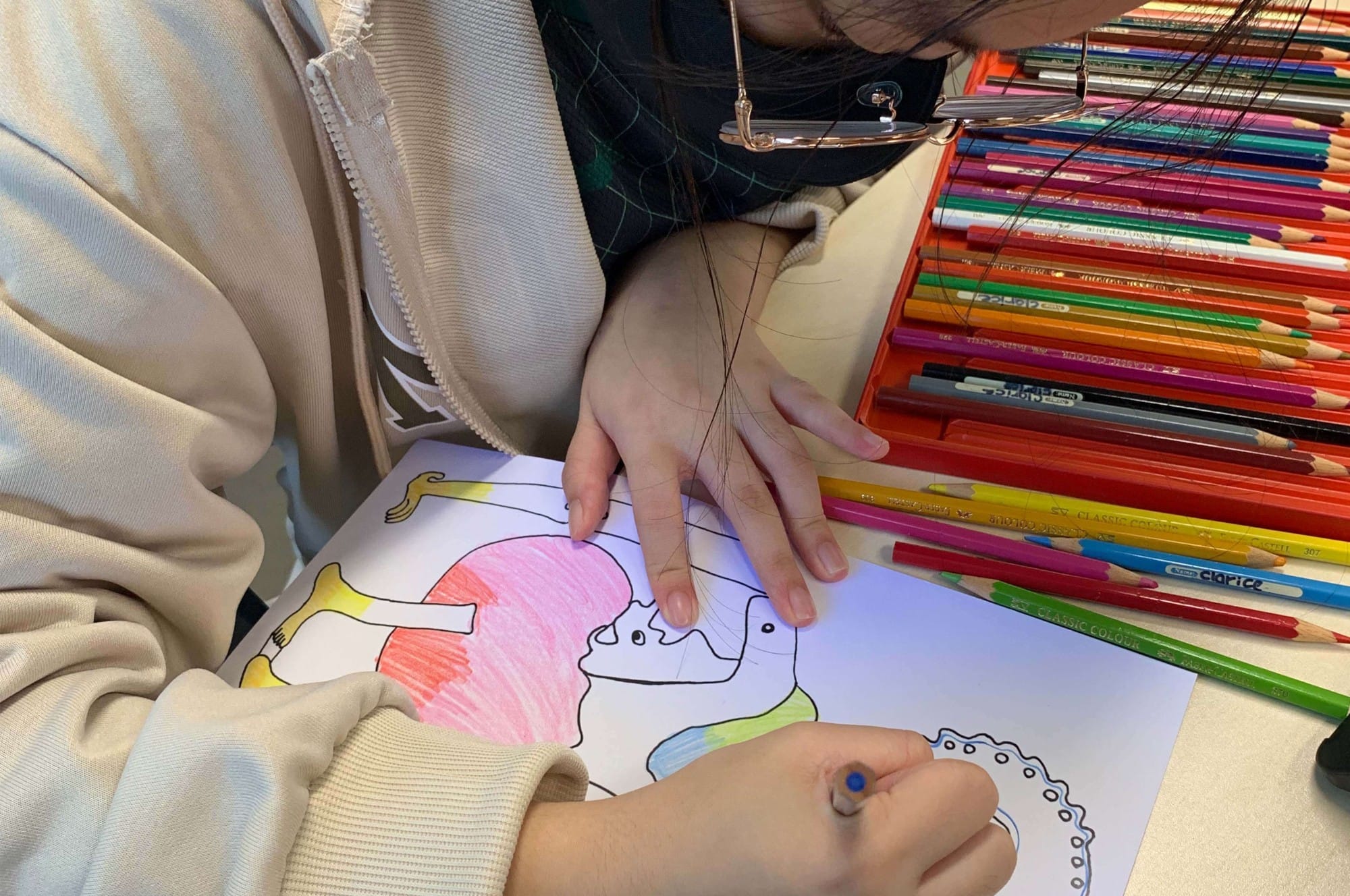 Person coloring a cartoon cat drawing with colored pencils, a large set of pencils arranged nearby.