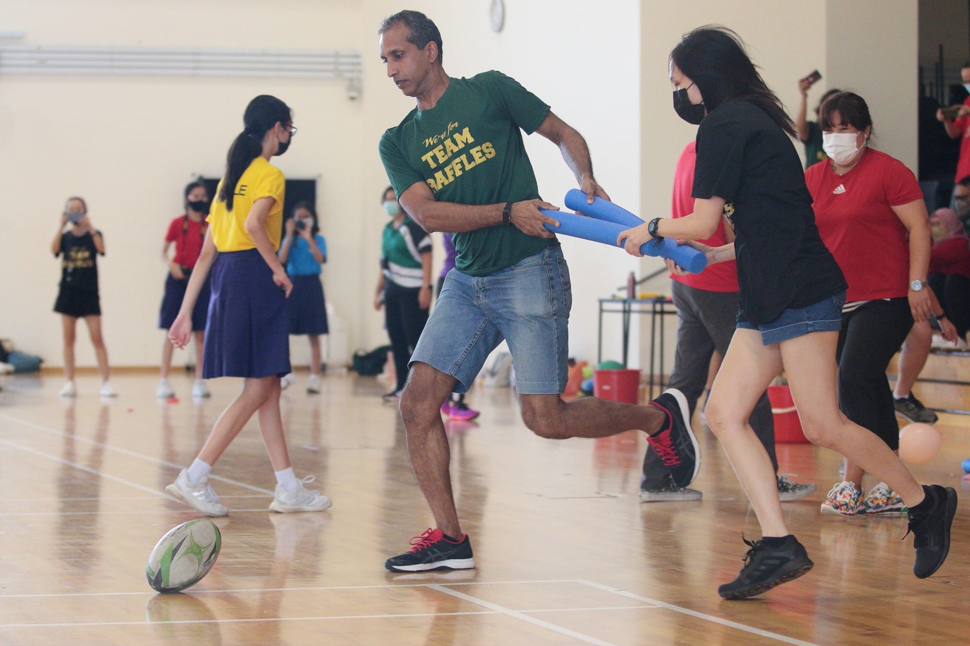 Two people pass blue foam batons in an indoor gym activity while others watch and a rugby ball sits on the floor.