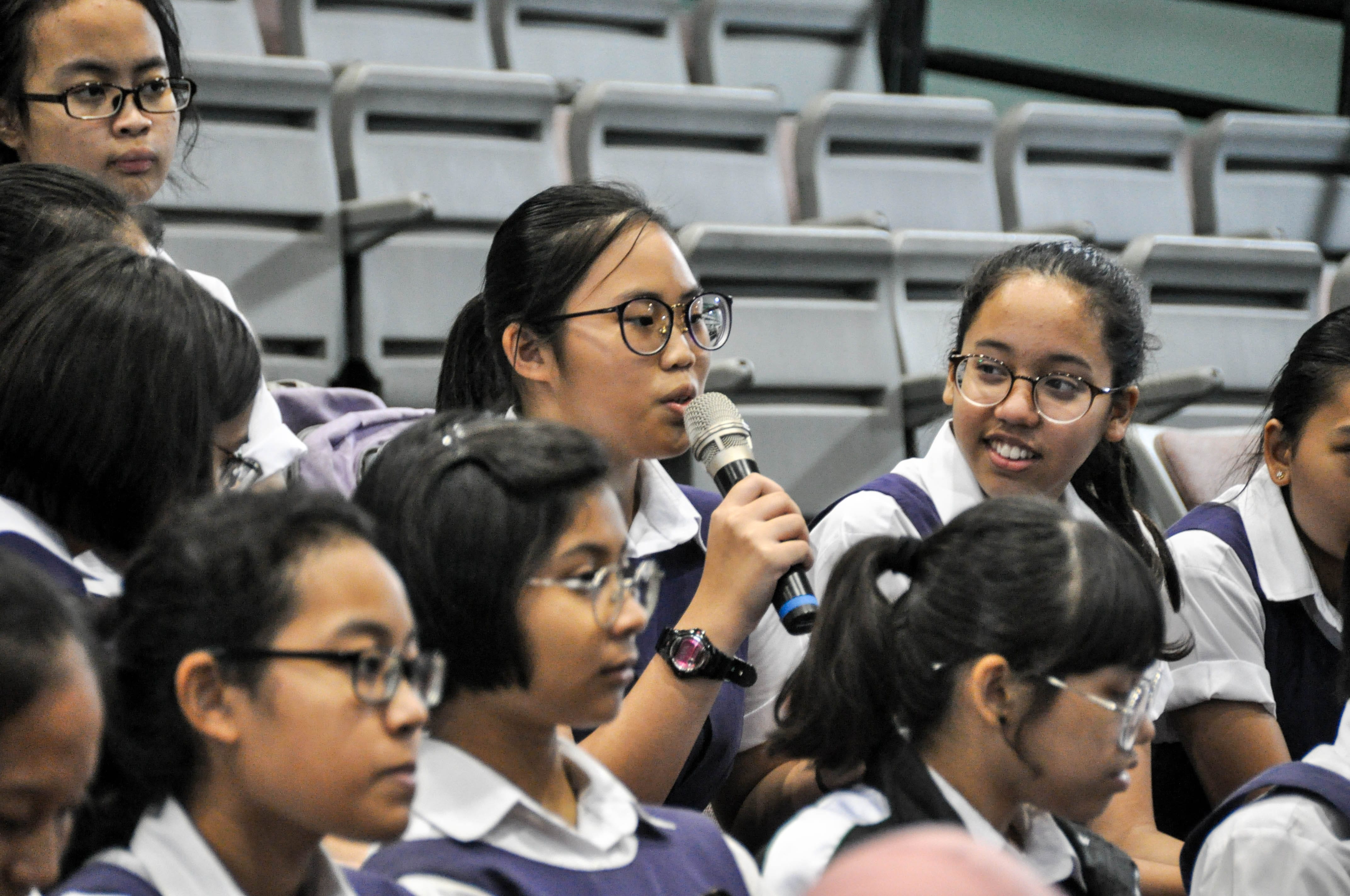 A young student in school uniform speaks into a microphone while seated among peers in an auditorium.