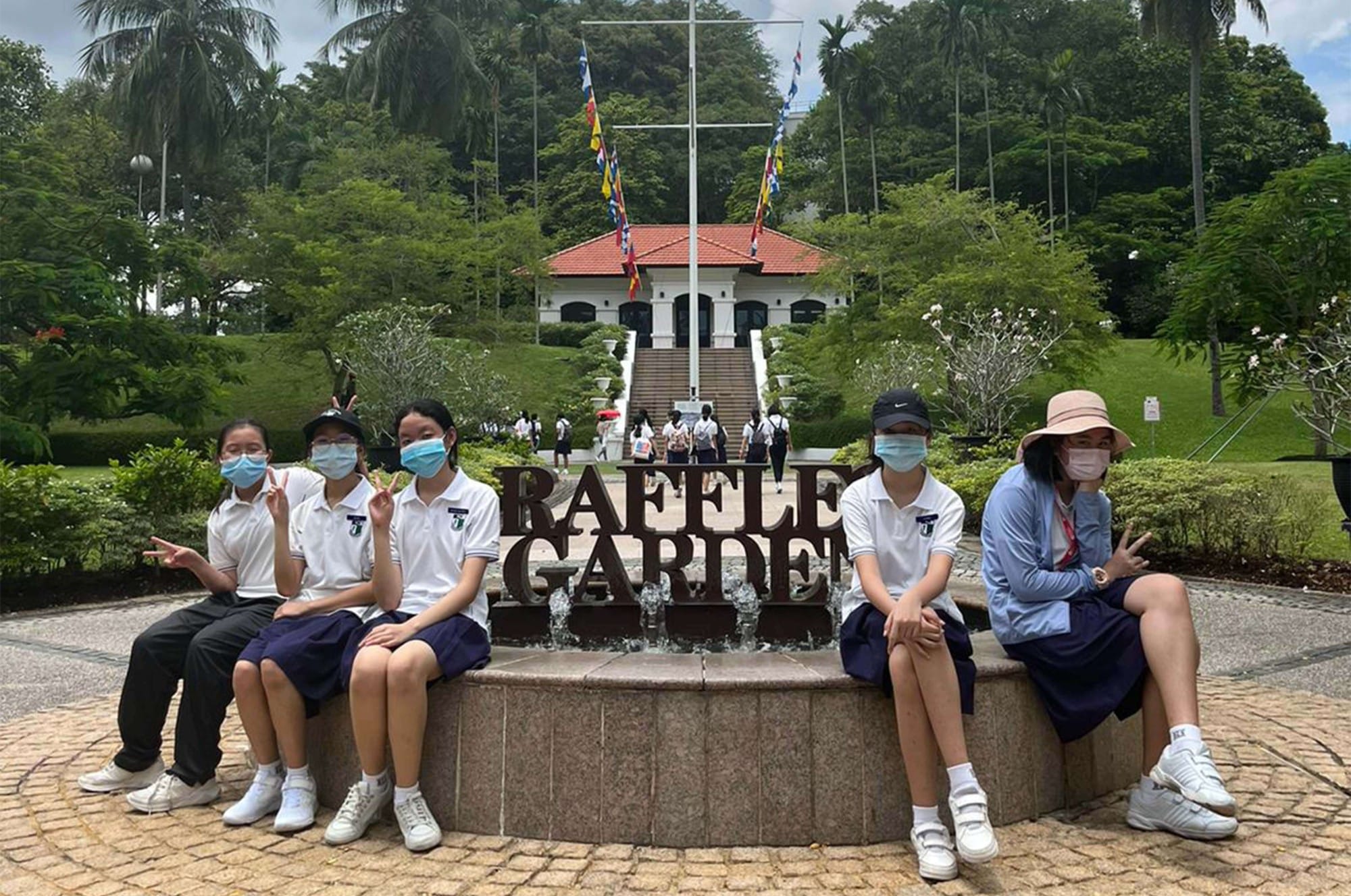 Five masked students in white uniforms sit on a fountain edge at Raffles Garden, with a white colonial building behind them.