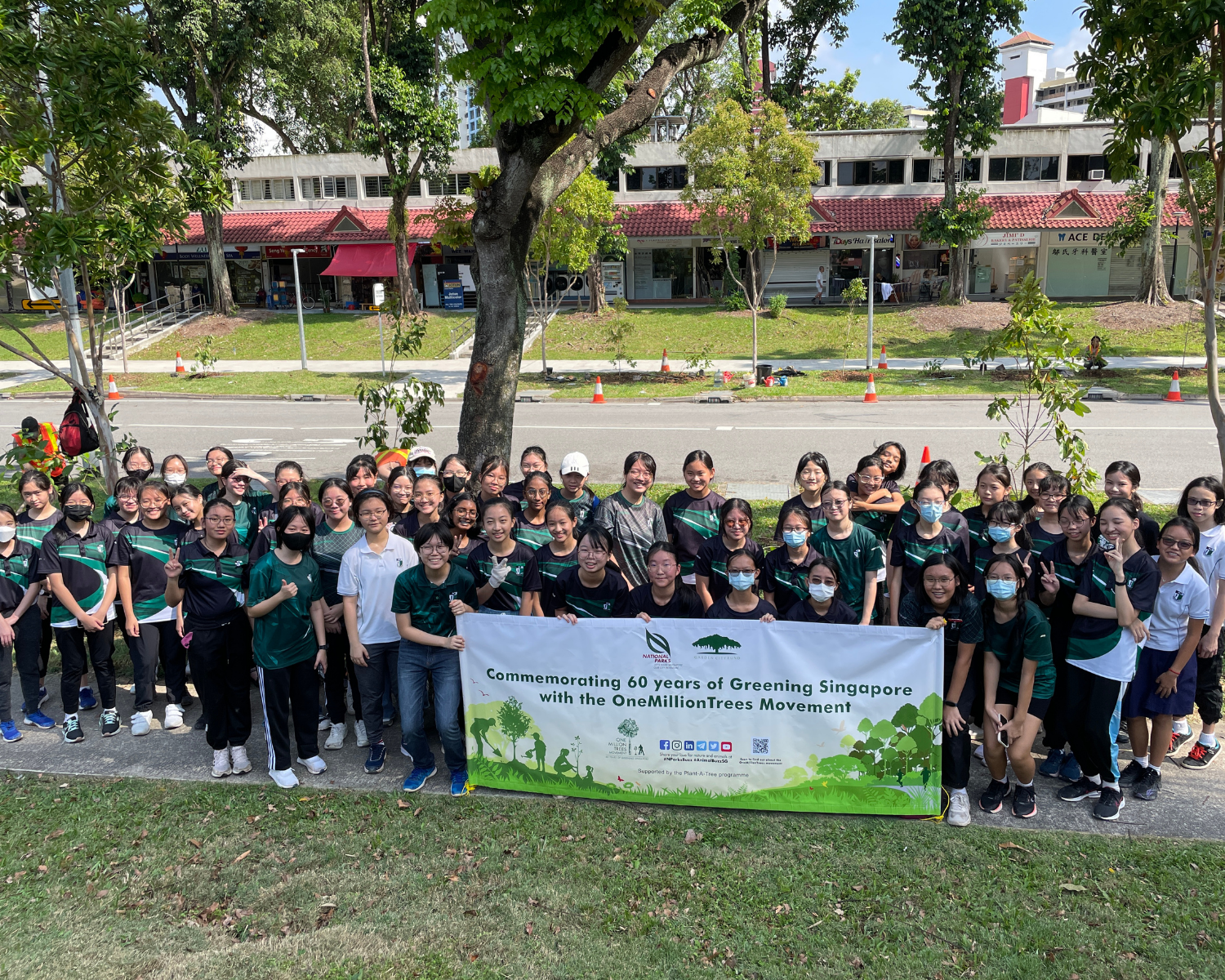 Large group holding a Green Singapore banner, standing on grass by a road under a big tree.