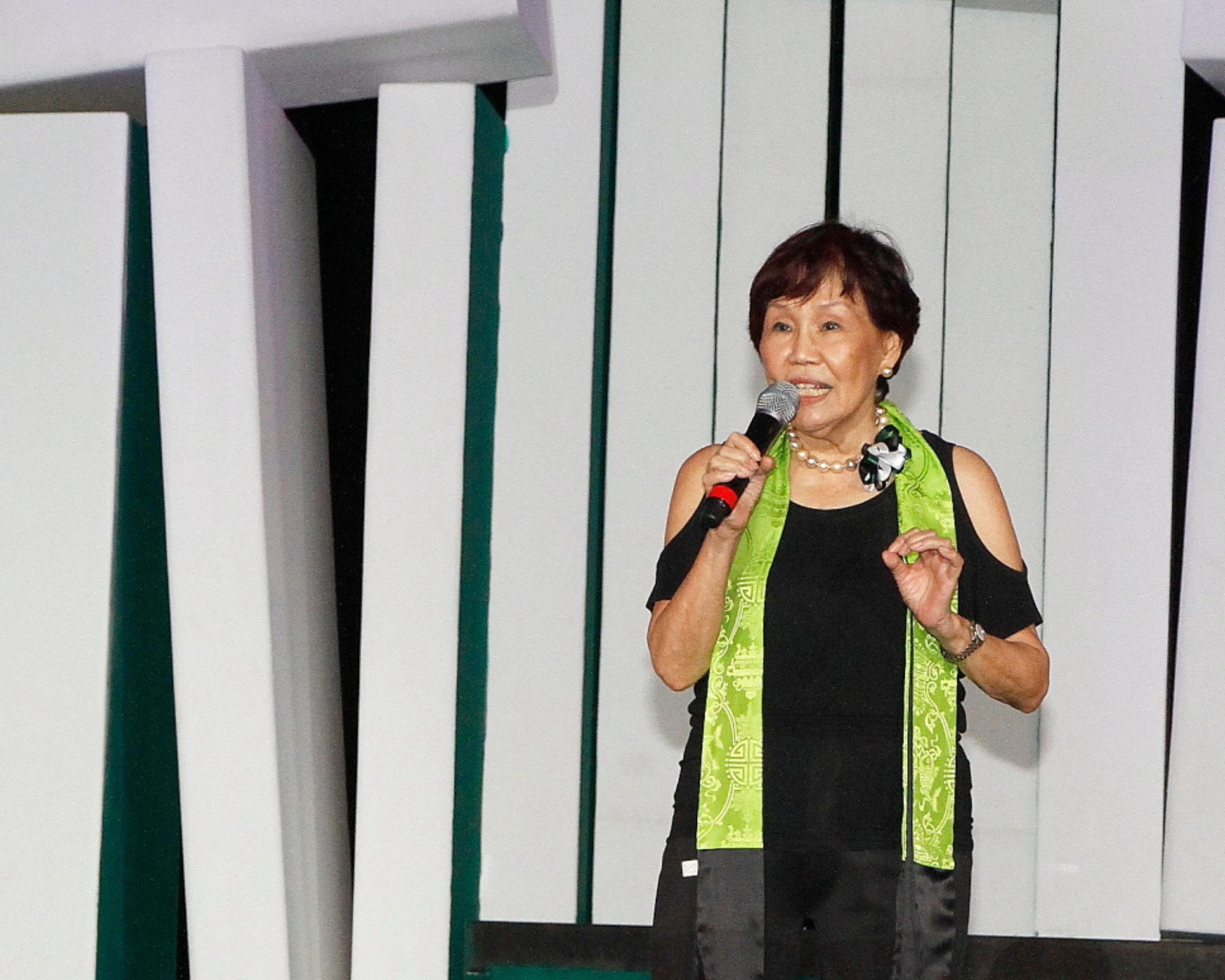 Elderly woman in black top with green scarf speaks into a microphone on a stage with white and green panels.