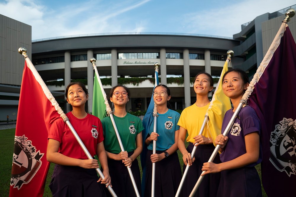 Five young girls in colorful school uniforms hold matching flags outdoors in front of a large circular building.