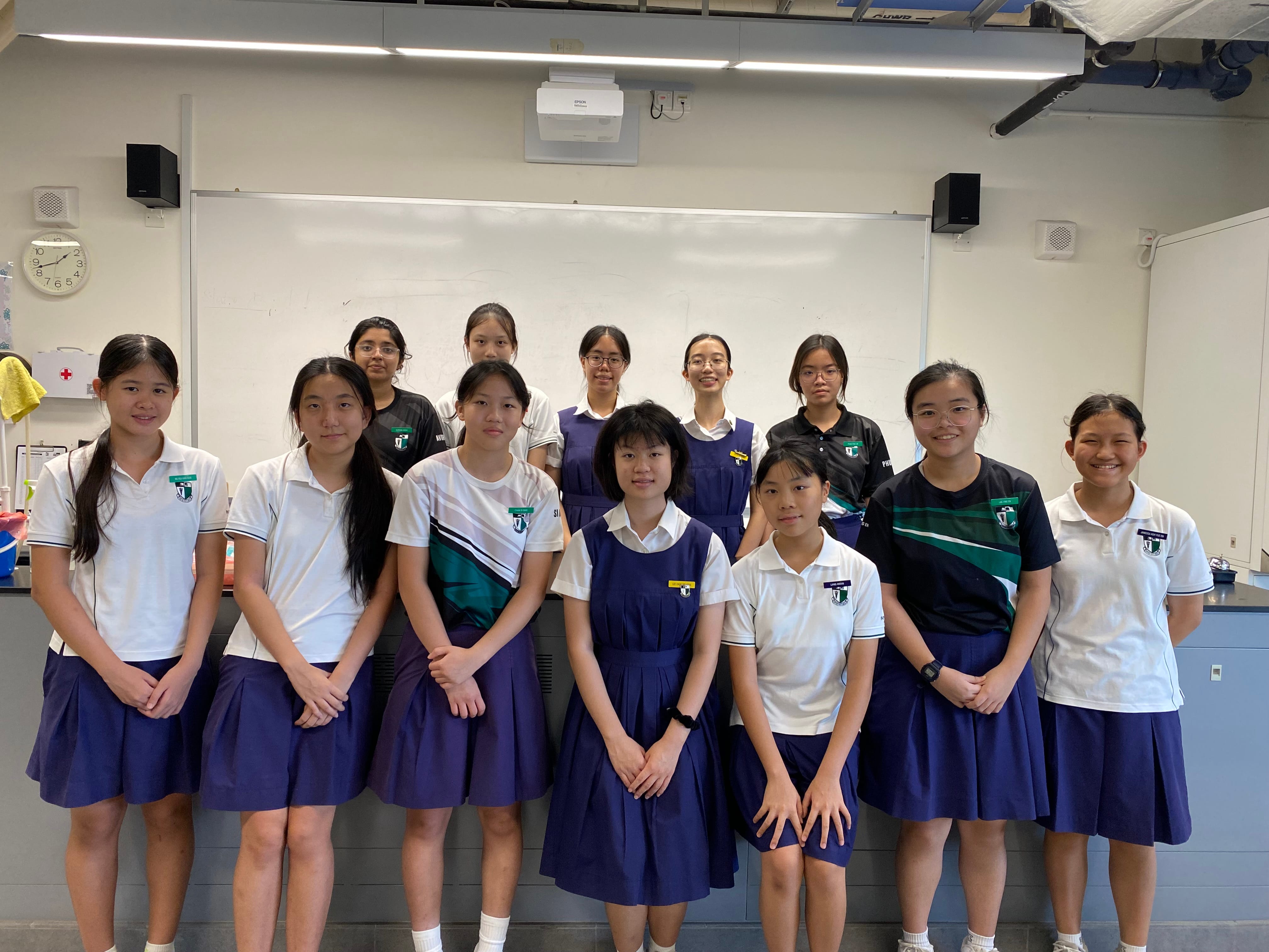 A group photo of female students in navy uniforms and white polo tops, standing in a science lab gathered together for a picture.