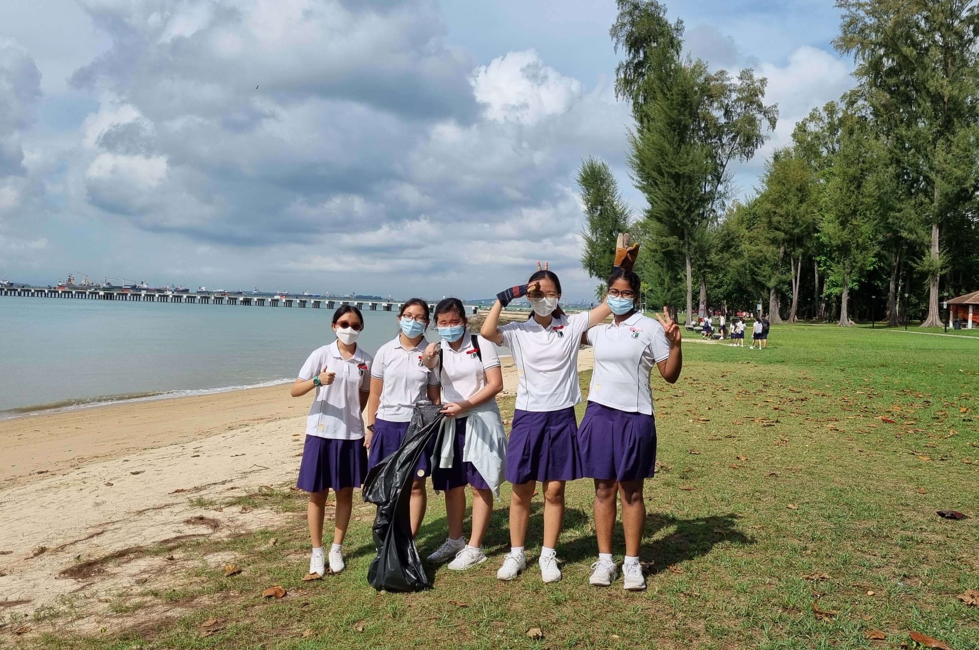 Five masked schoolgirls in white and purple uniforms pose on a beach holding a trash bag during a cleanup.