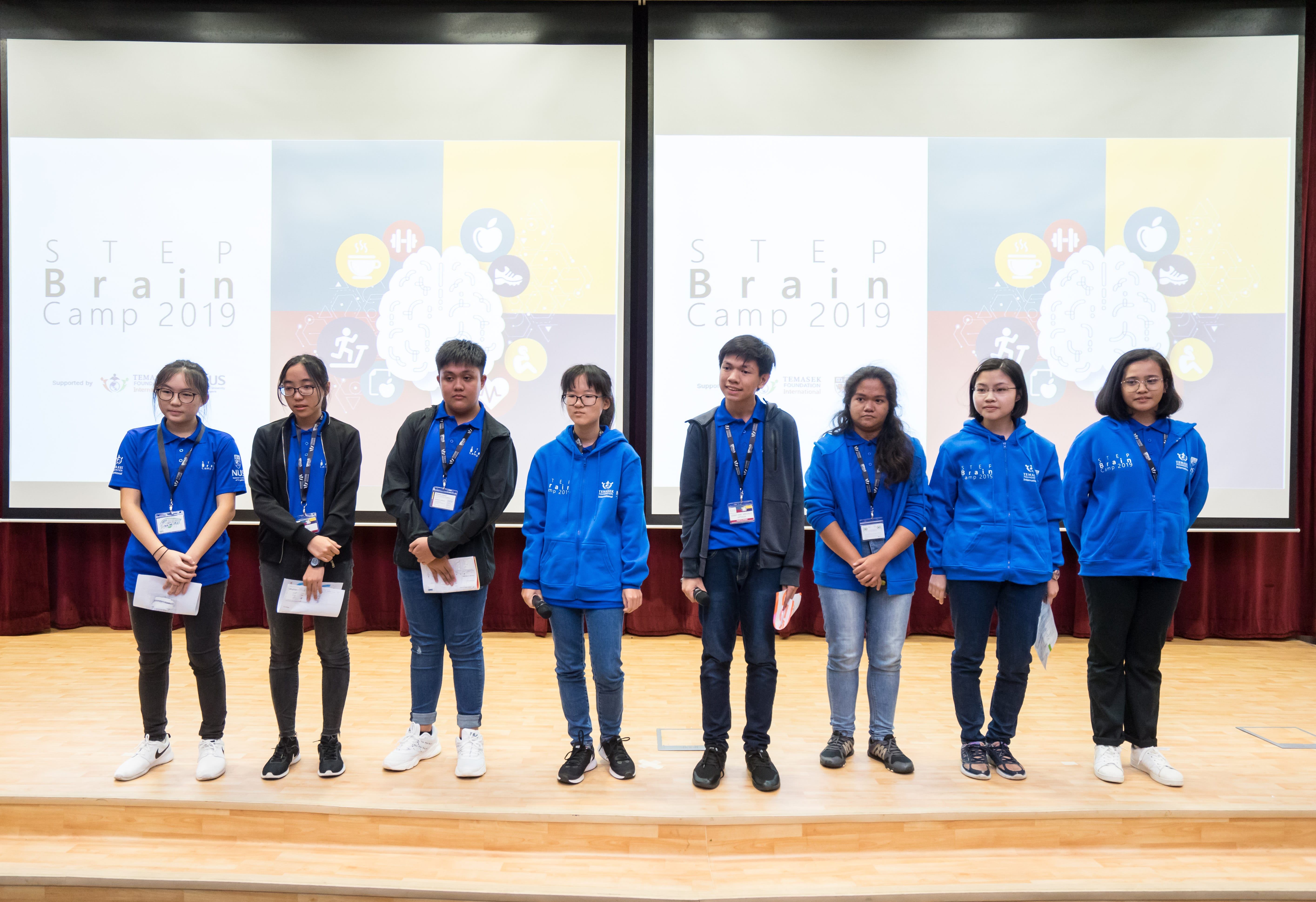 Eight young students in blue hoodies stand on a stage in front of screens displaying STEP Brain Camp 2019.