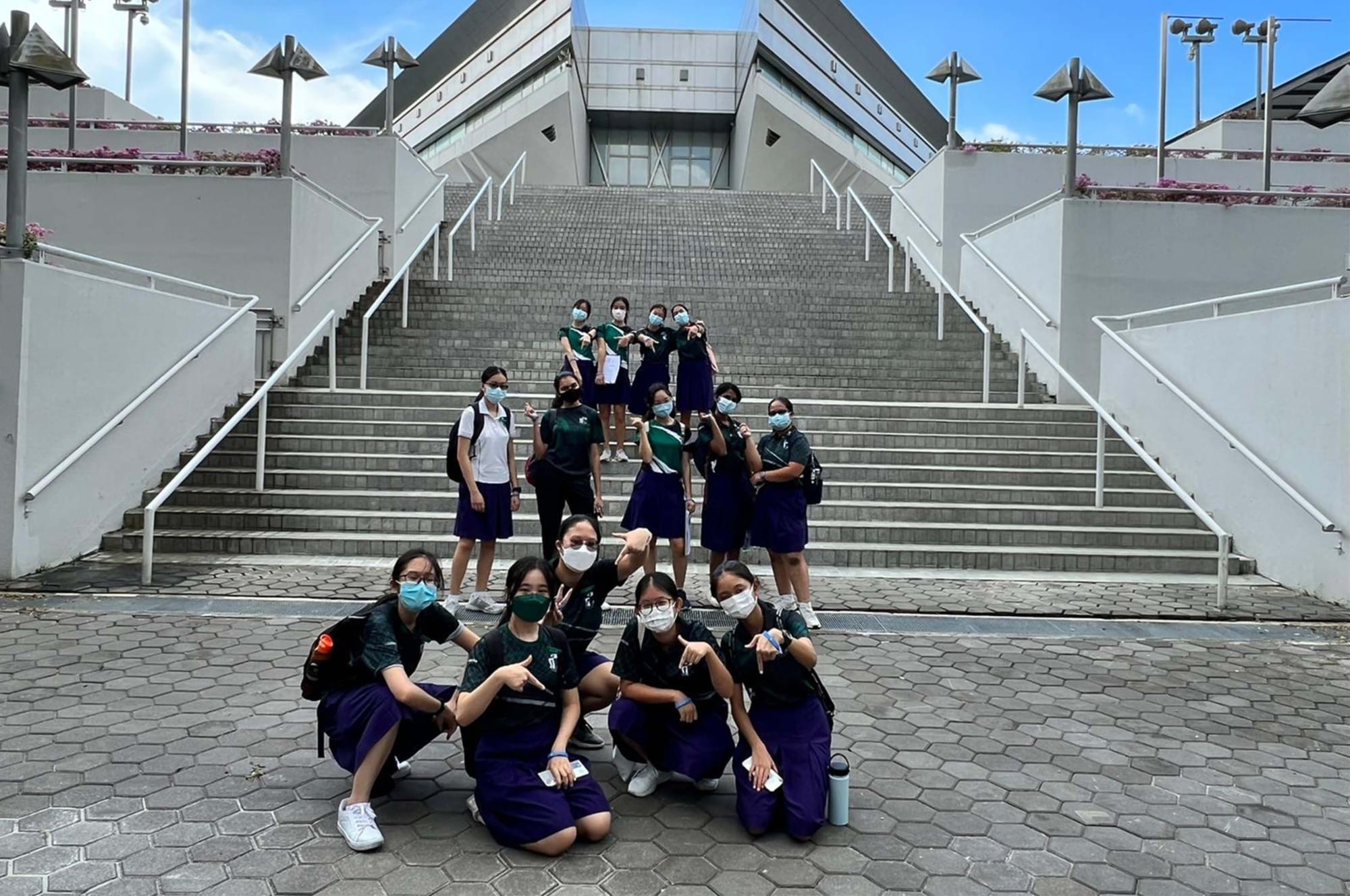 A group of masked female students in navy uniforms pose on wide outdoor stairs in front of a modern building.