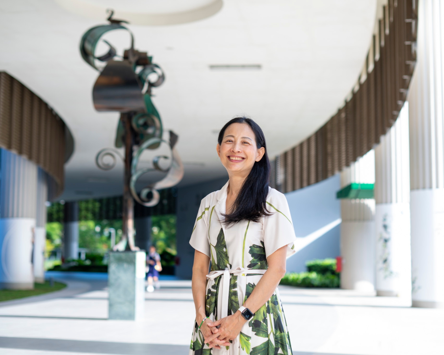 A smiling woman in a floral dress stands under a curved white overhang with a metal sculpture behind her.