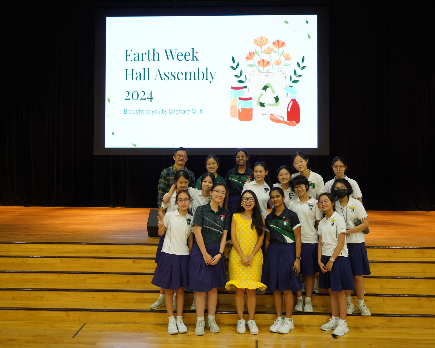 Group of students and a teacher posing on stage in front of an Earth Week Hall Assembly 2024 presentation slide.