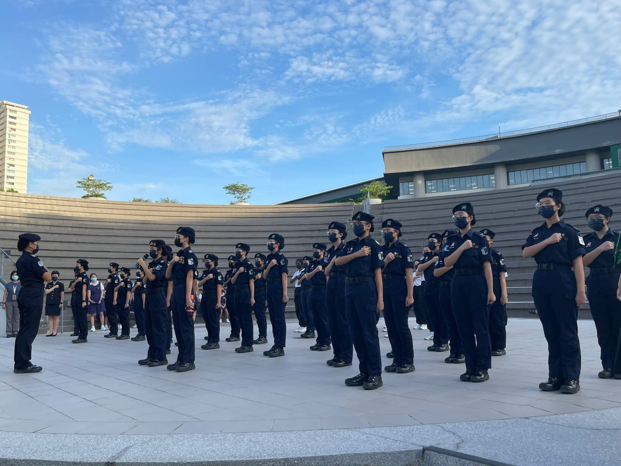 Rows of uniformed cadets in dark outfits and berets stand at attention outdoors with fists raised to their chests.