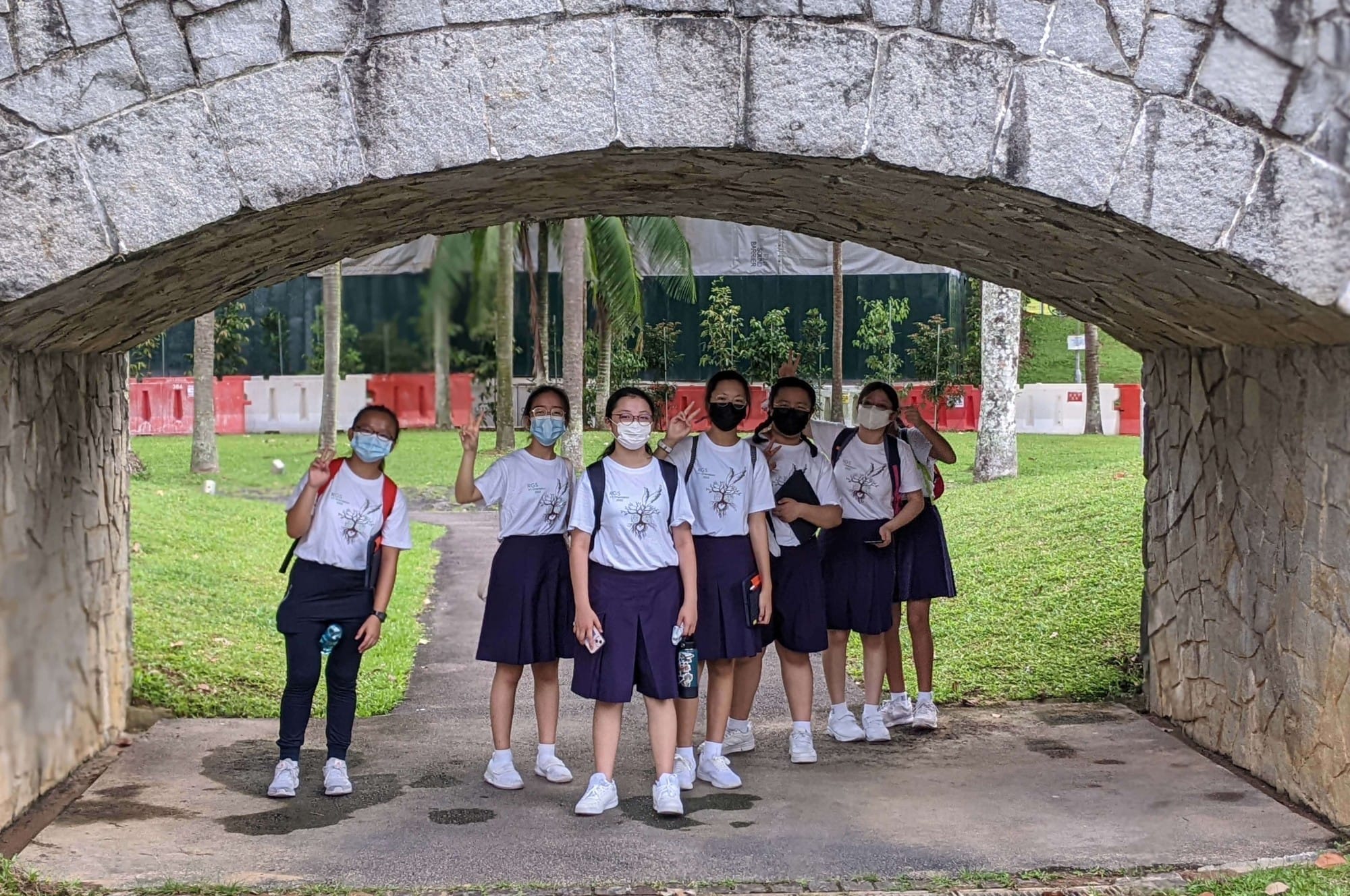 Six masked schoolgirls in white shirts and navy skirts pose under a stone arch in a green park.