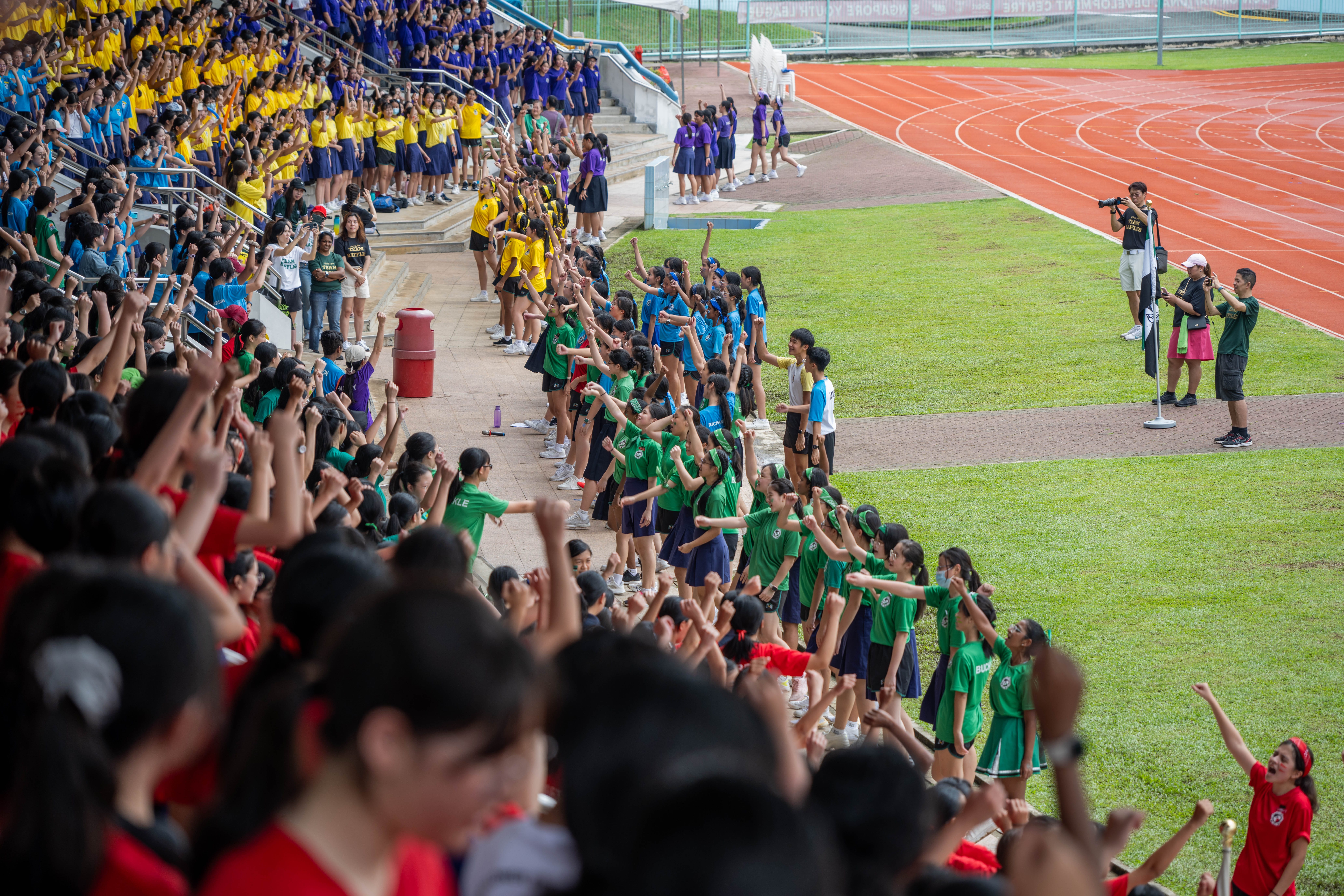 Crowds in colorful shirts cheer in stadium stands beside a running track, arms raised in unison.