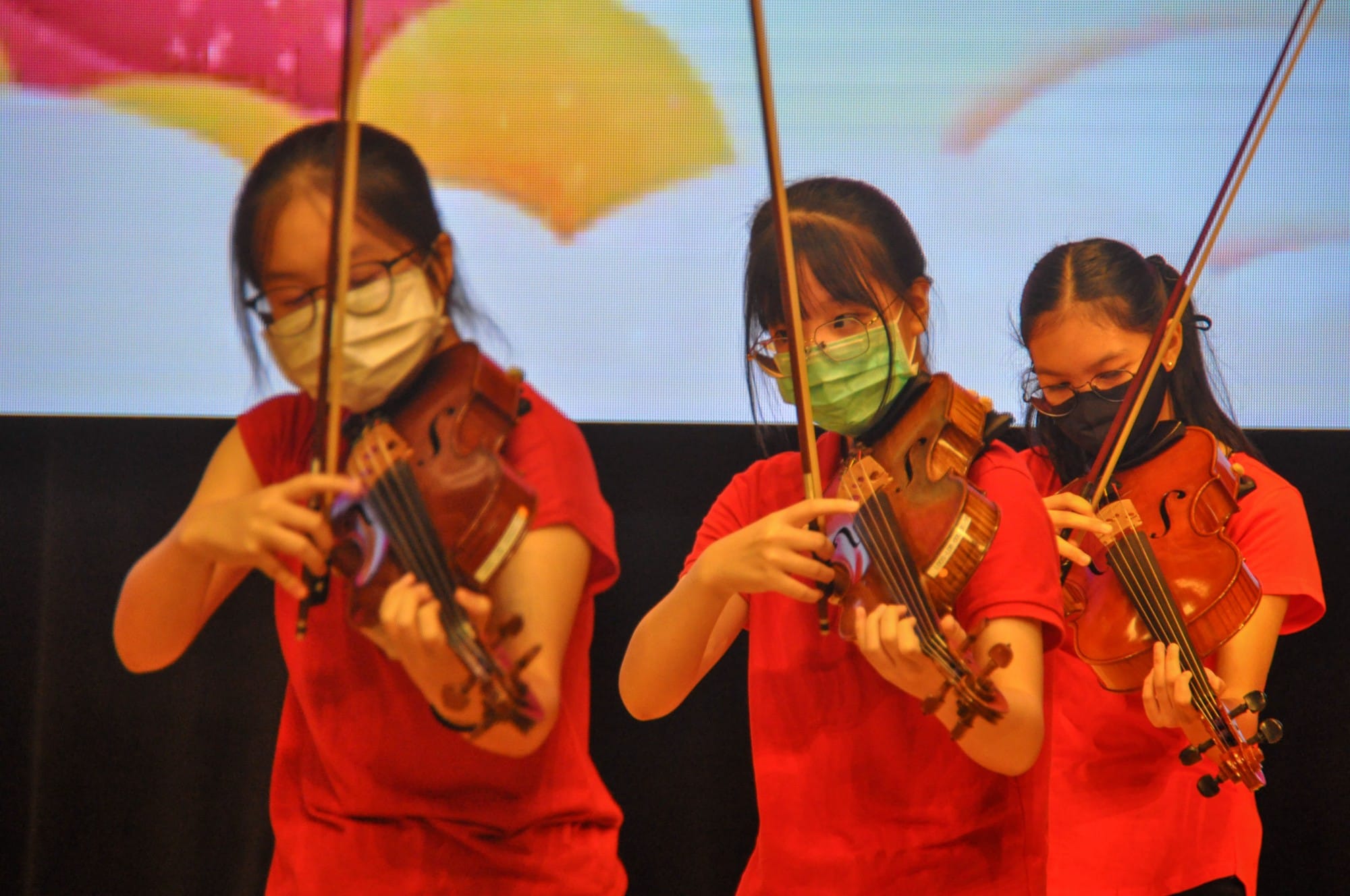 Three young girls in red shirts and face masks play violins on stage before a colorful floral backdrop.