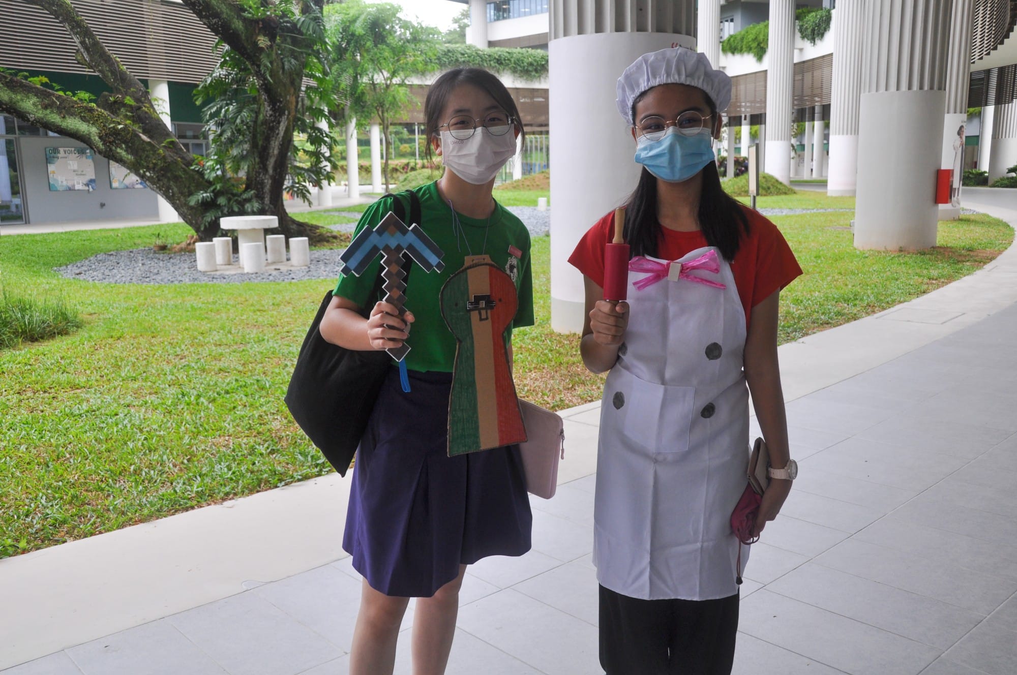 Two masked girls in costumes stand outdoors; one holds a Minecraft pickaxe, the other wears a chef outfit with a rolling pin.