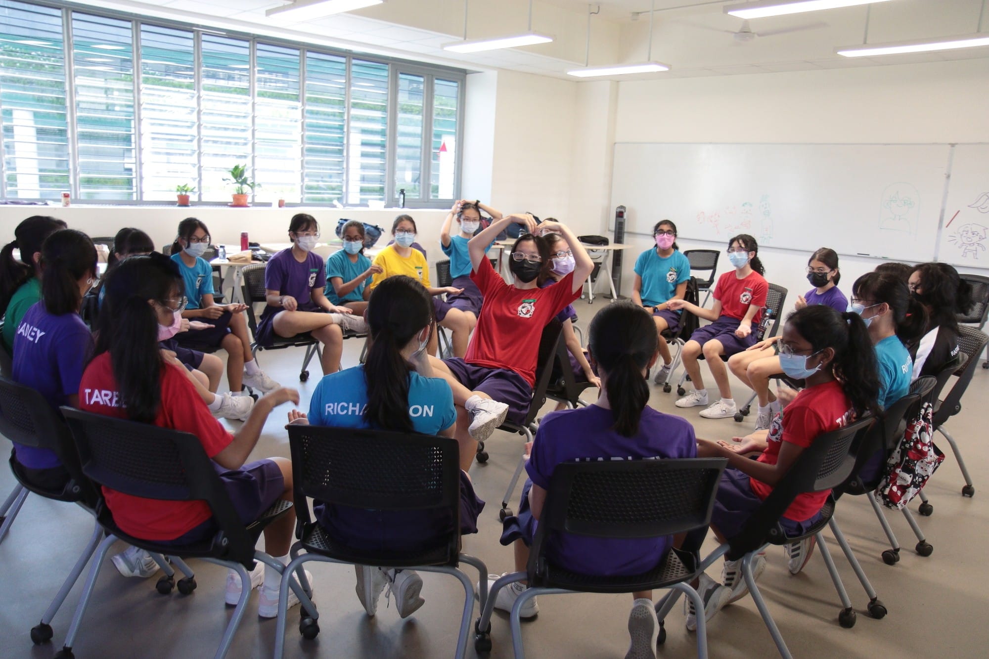 Students in colorful uniforms and face masks sit in a circle in a bright classroom during a group activity.