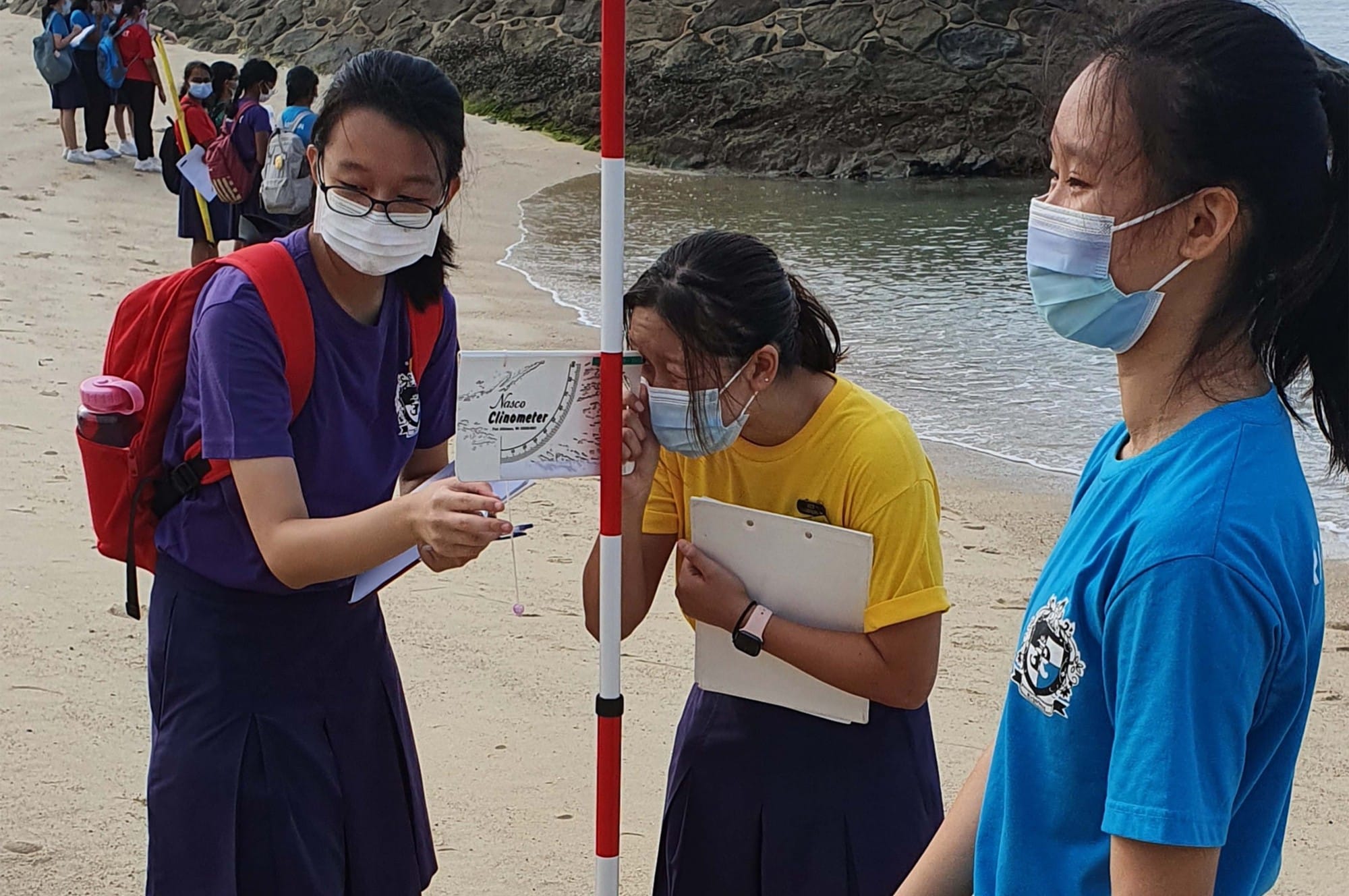 Three masked students use a clinometer and survey pole on a sandy beach during an outdoor field study.