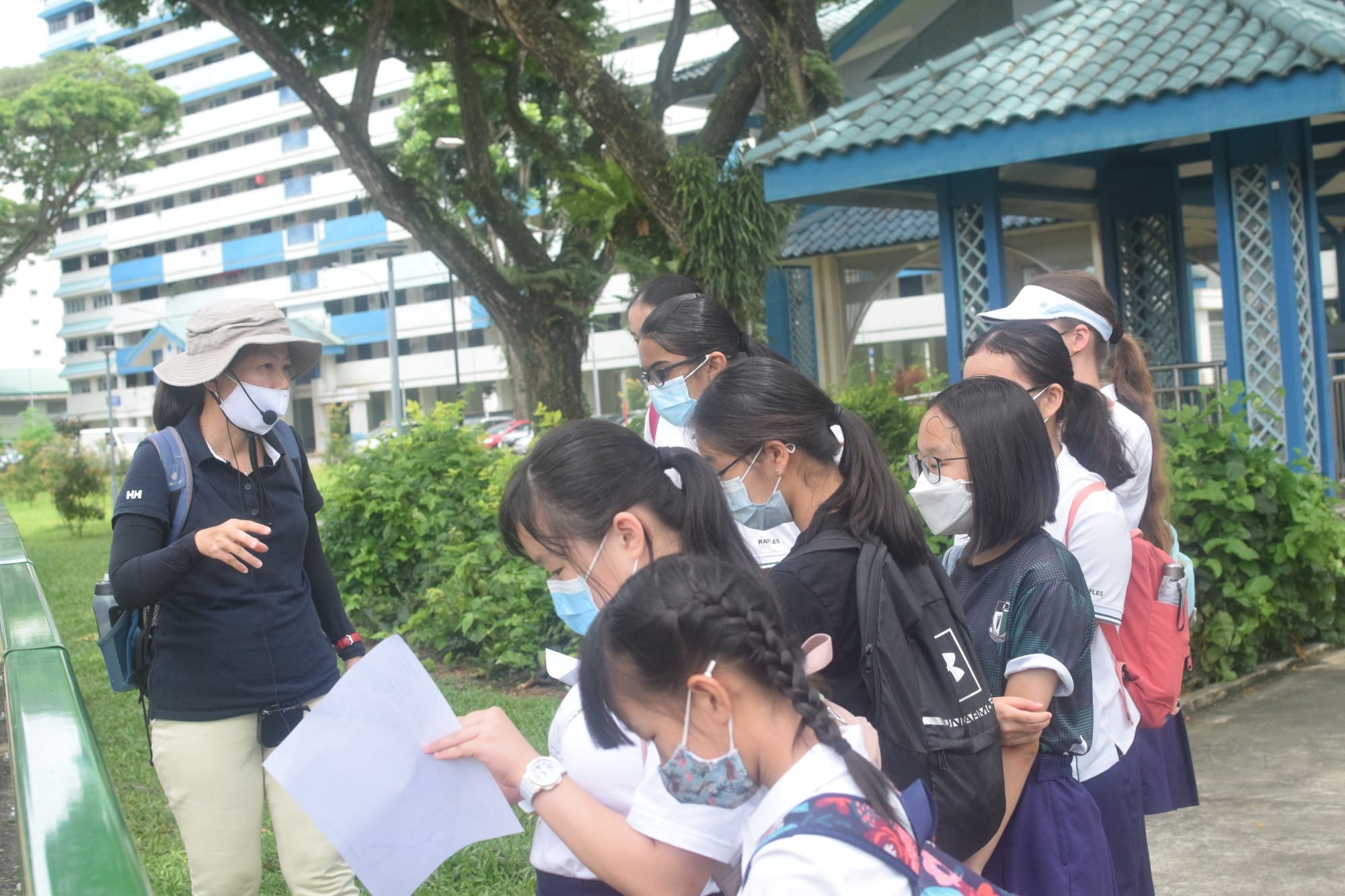 A masked adult guide speaks to a group of masked students holding papers outdoors near a blue building.