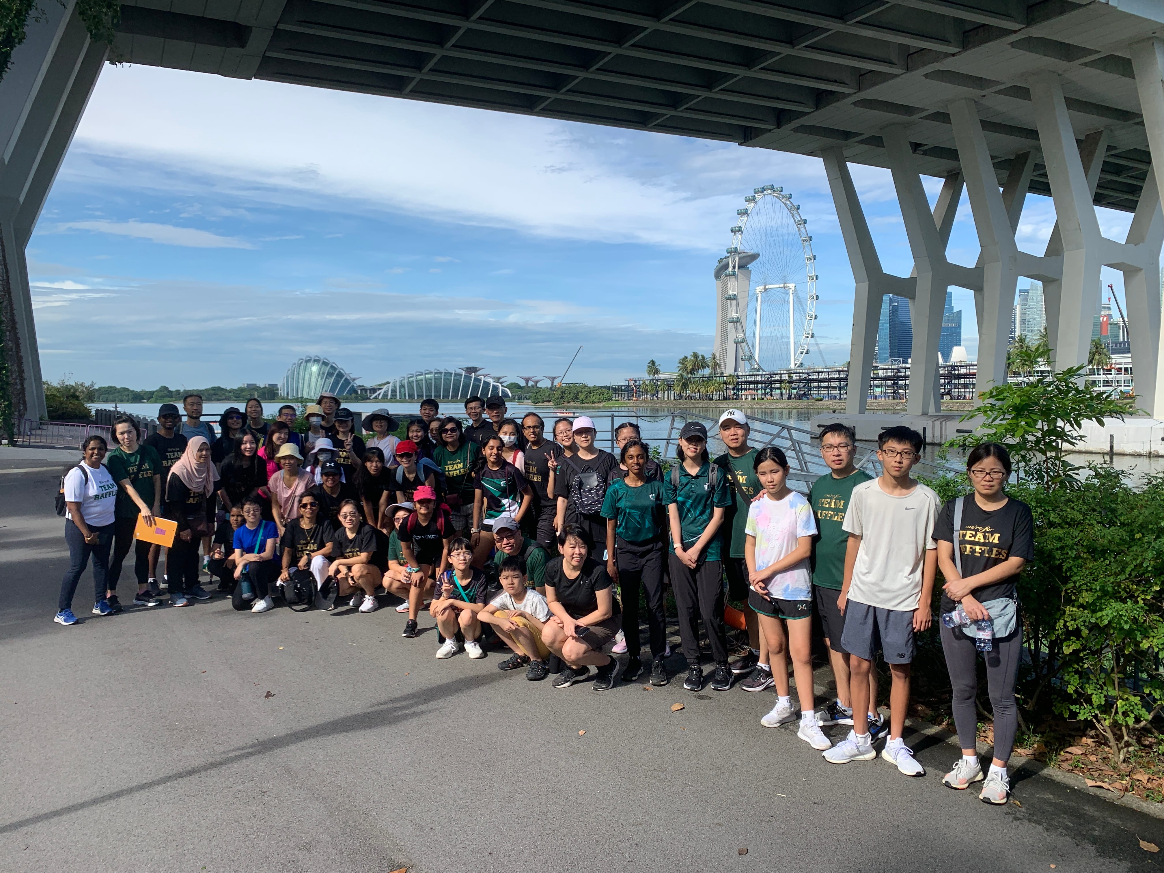 Large group posing under a bridge by the water, with a giant Ferris wheel and skyline in the background.