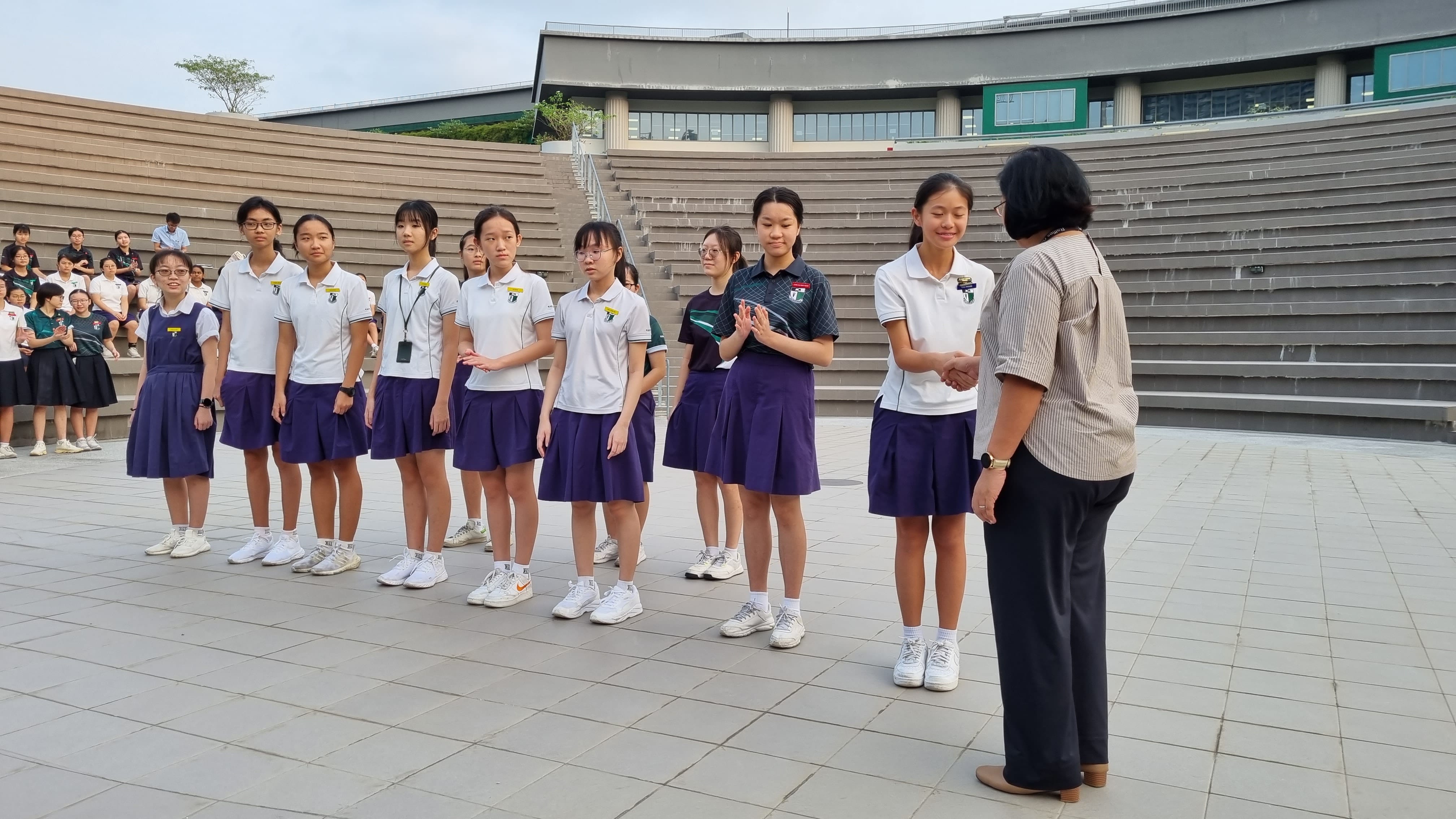 A row of female students in white polo shirts and purple school uniform skirts stand outdoors as an adult shakes one student's hand.