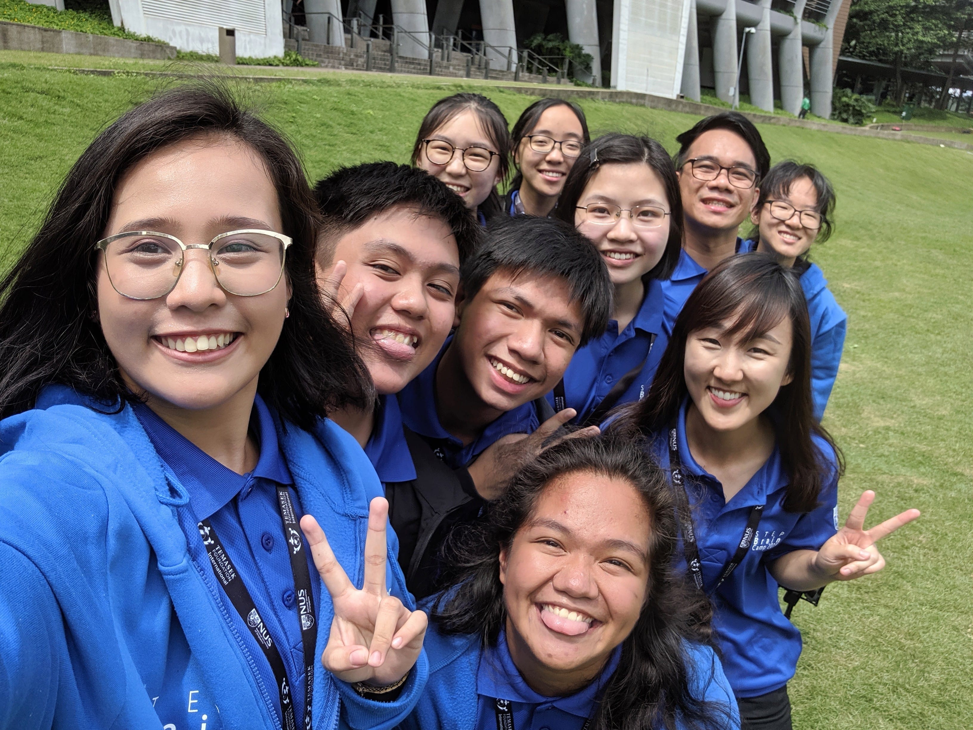 A group of smiling young people in matching blue shirts take a selfie outdoors on a green lawn near a building.