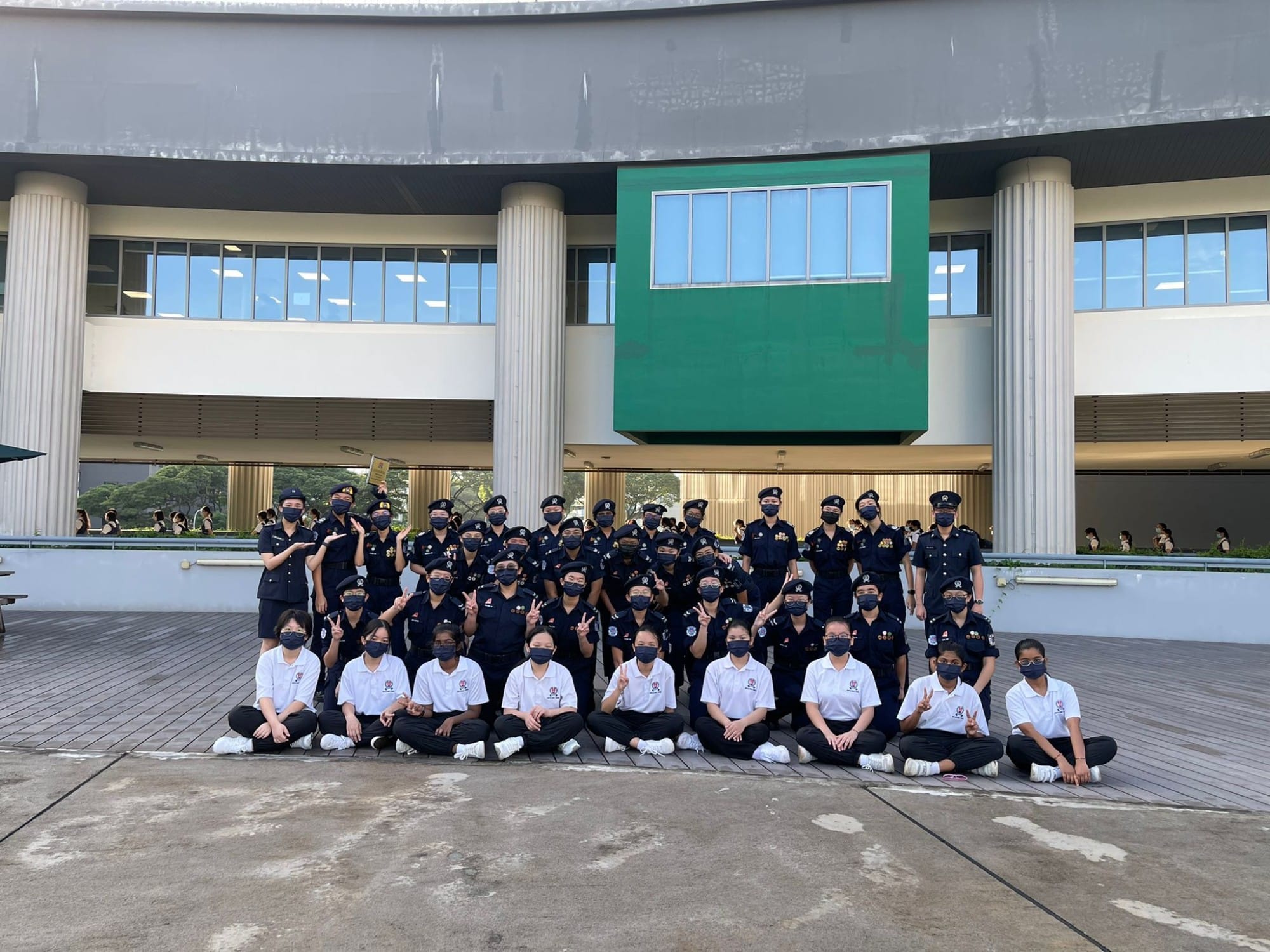 Large group of masked youth in dark uniforms and white shirts posing outdoors in front of a curved building.